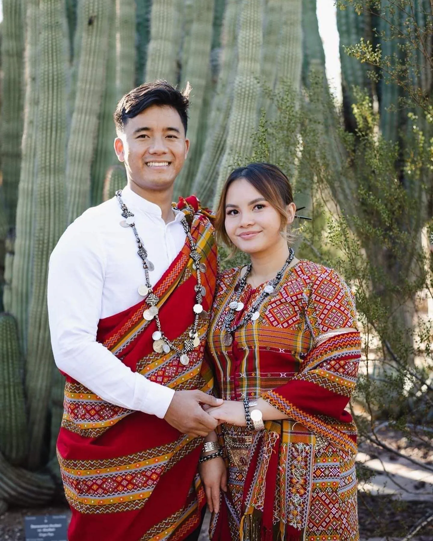 A smiling couple wearing traditional, colorful, patterned clothing and jewelry, standing in front of large cacti in a desert landscape.