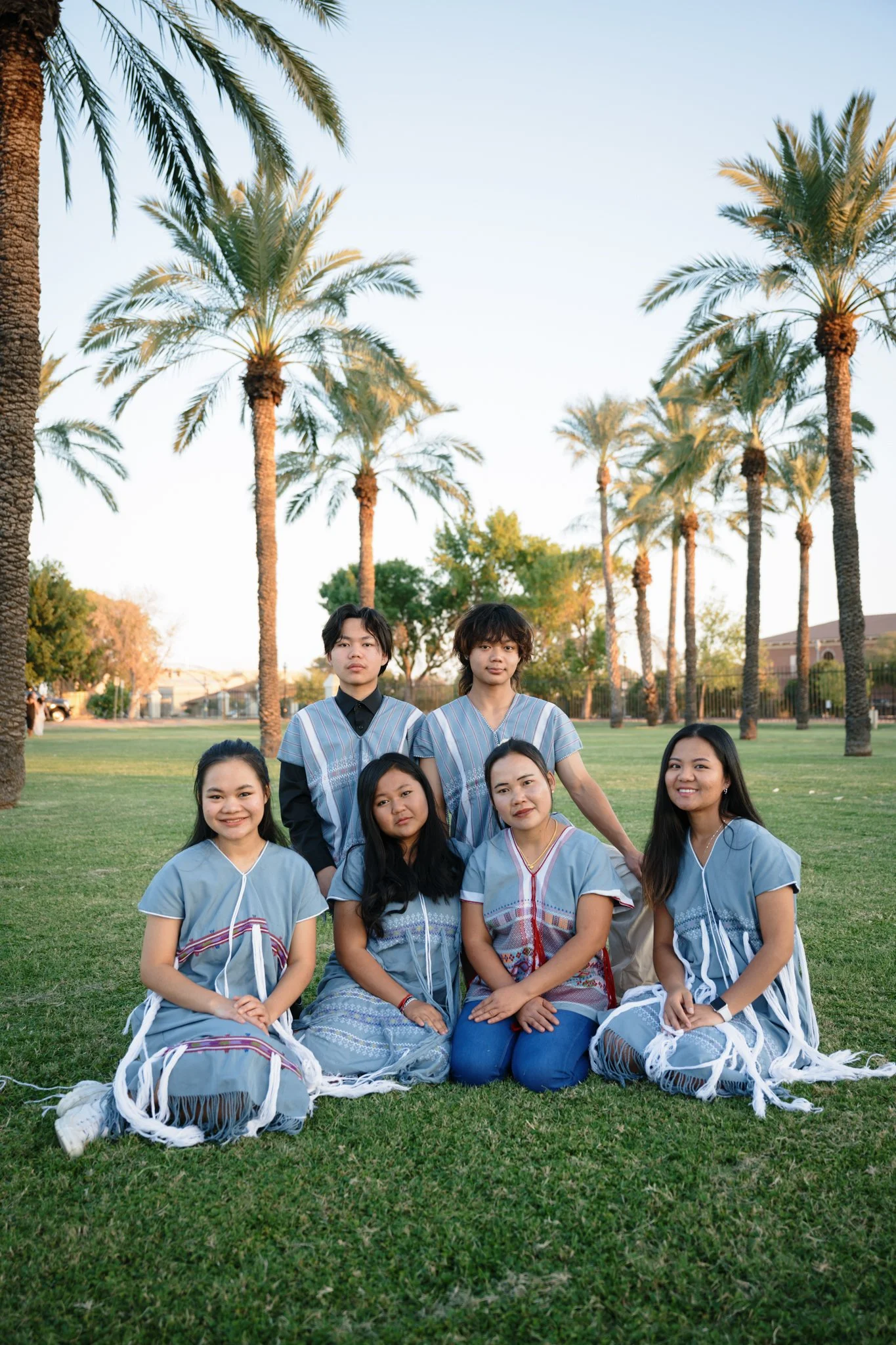 Group of seven young people, four women and three men, posing outdoors on a grassy area with tall palm trees in the background during late afternoon or early evening.