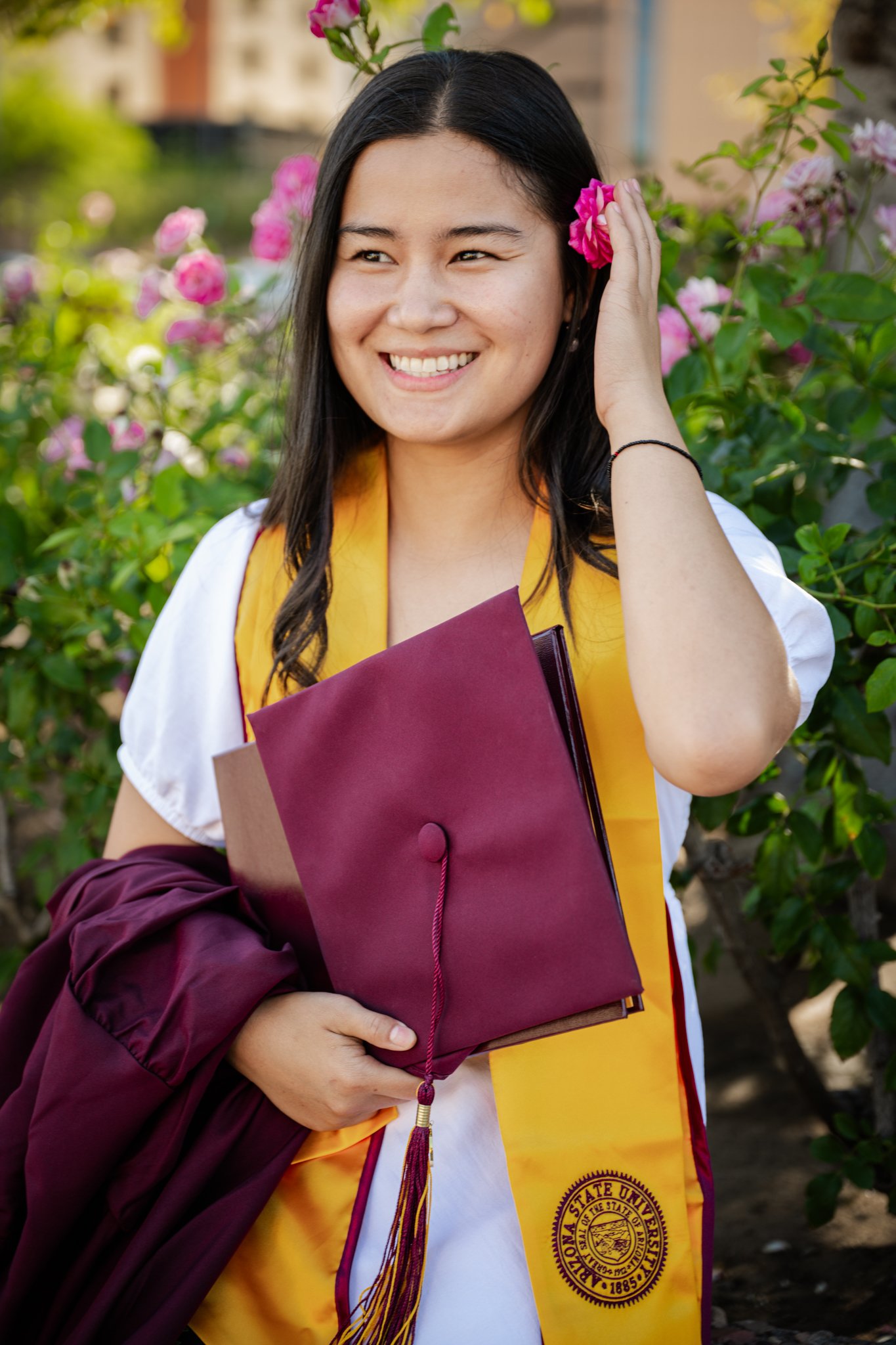 A young woman in graduation attire standing outdoors among pink flowers, holding a maroon diploma cover, smiling, and adjusting her hair.