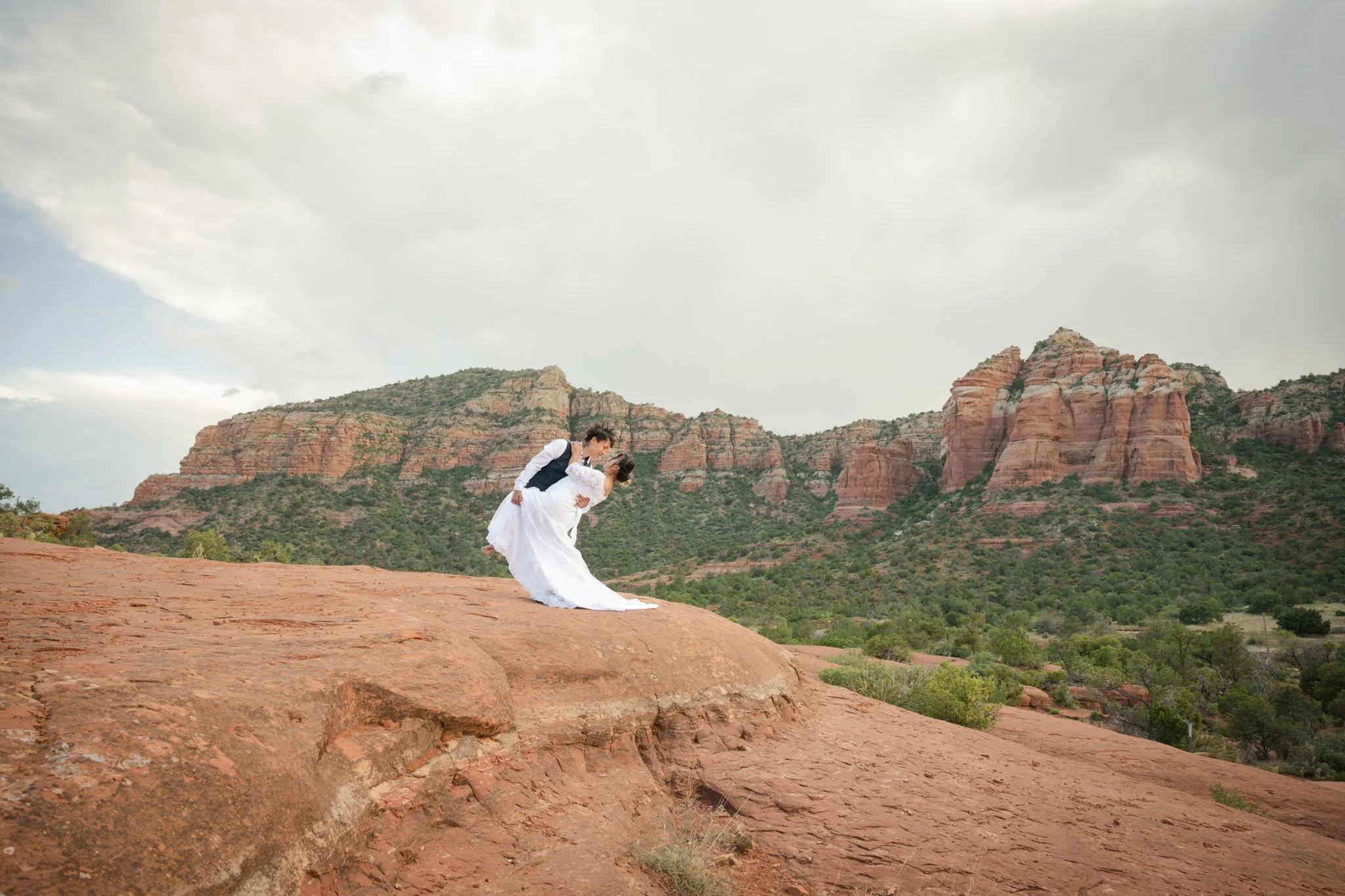 A bride in a white wedding dress being lifted by a groom in a vest and shirt on red rock formation with green vegetation and rocky hills in the background, under a cloudy sky.