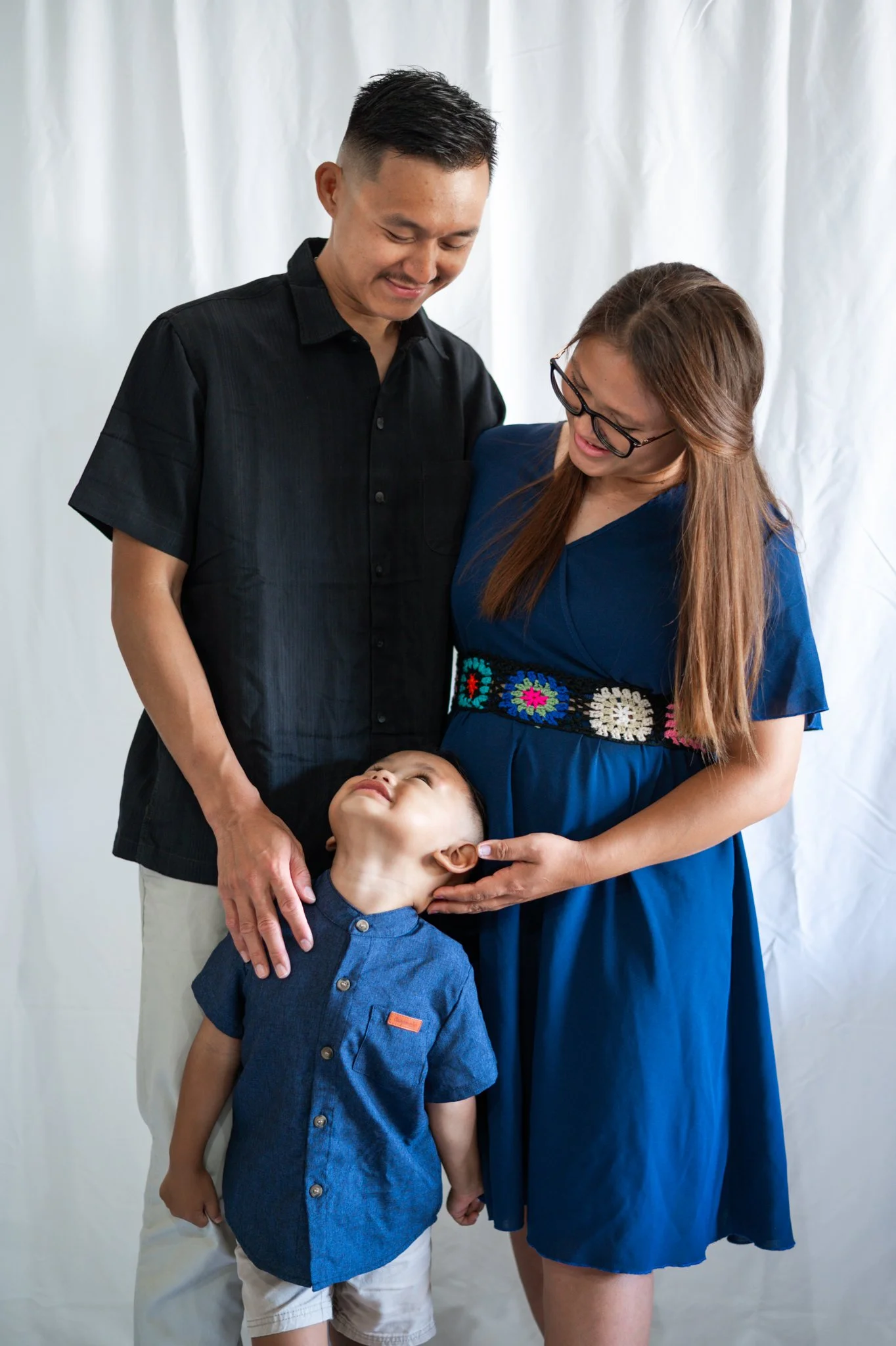 A family of four includes a woman in a blue dress with a floral pattern around the waist and glasses, a man in a black shirt, a young boy in a denim shirt, and a young girl with glasses. The woman and man are smiling and looking down at the children, who are looking upward. The background is plain white.