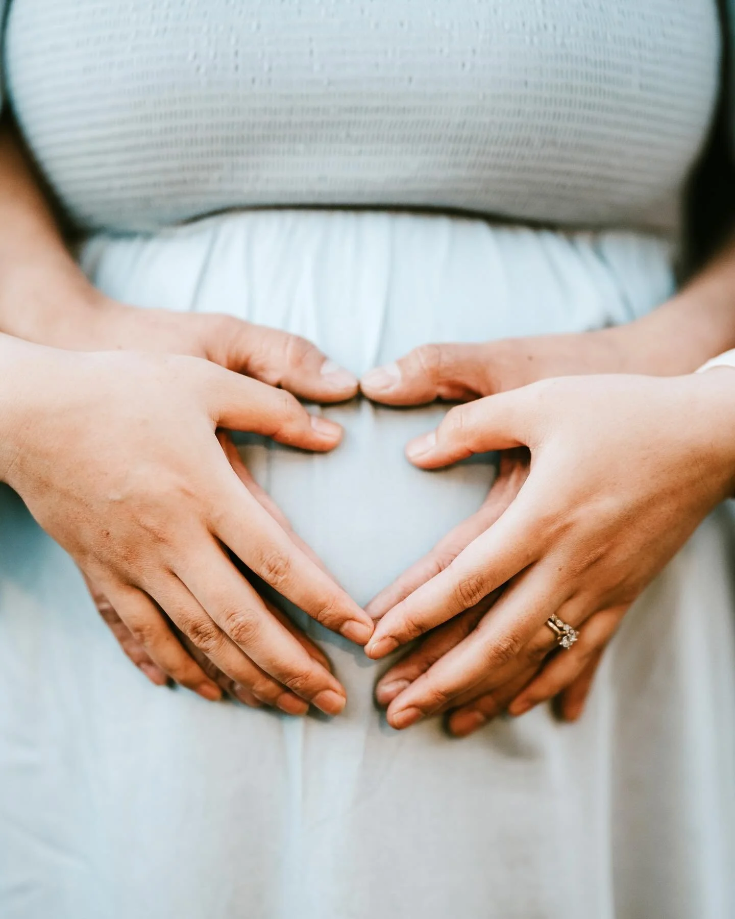 A person wearing a light-colored dress with hands forming a heart shape over their pregnant belly.