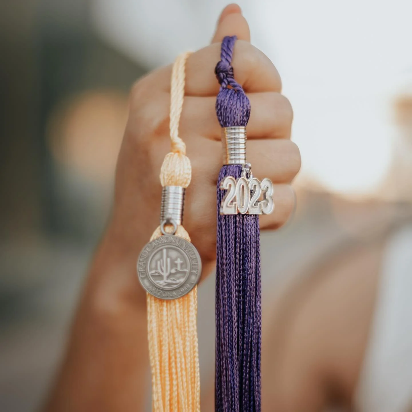 Close-up of a person's hand holding two graduation cords with medals and a purple tassel that has a silver charm with the year 2023, in front of a blurred background.