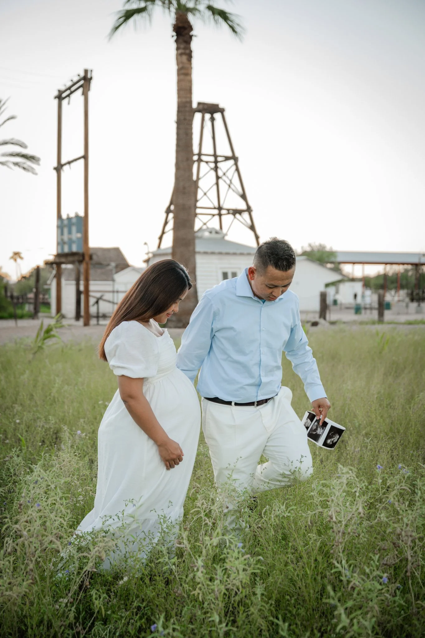 A pregnant woman and a man looking at ultrasound images while standing in a grassy field with palm trees, buildings, and an old water tower in the background.