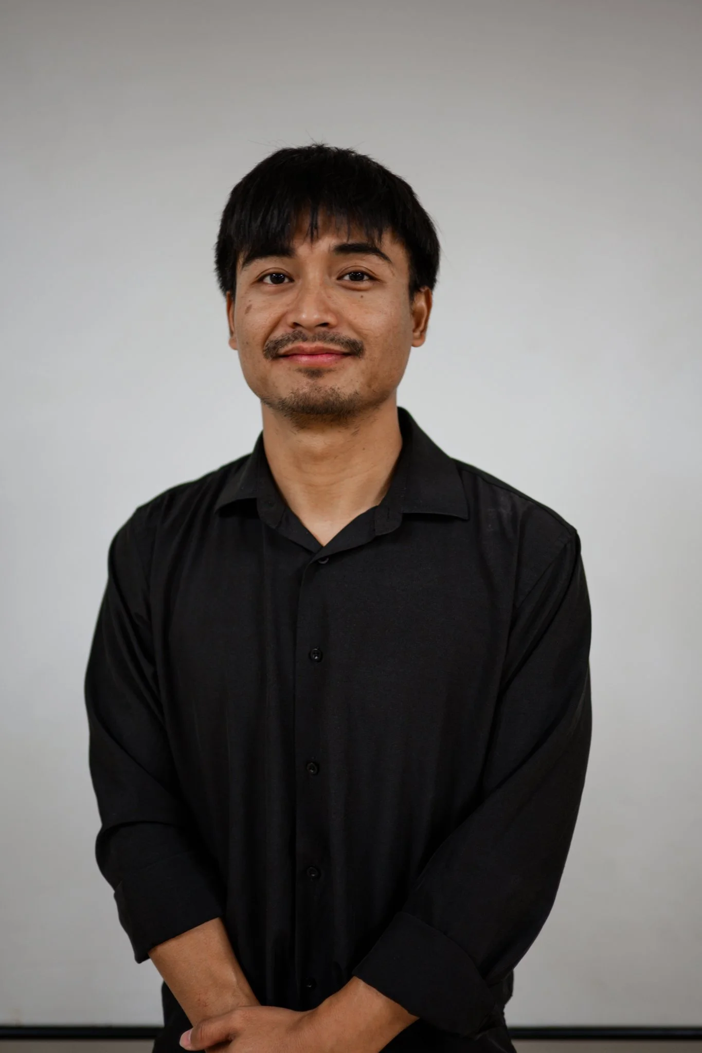 A man with short black hair, a slight mustache, and a goatee, dressed in a black shirt, standing against a plain light gray background, looking at the camera with a slight smile.
