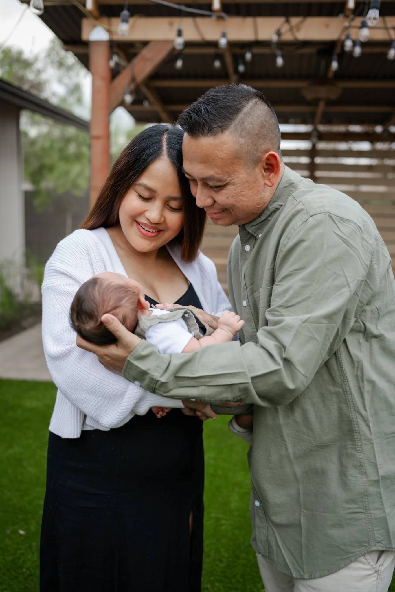 A couple holding and looking at a newborn baby outdoors under a wooden pergola with string lights, in a garden with green grass and trees.