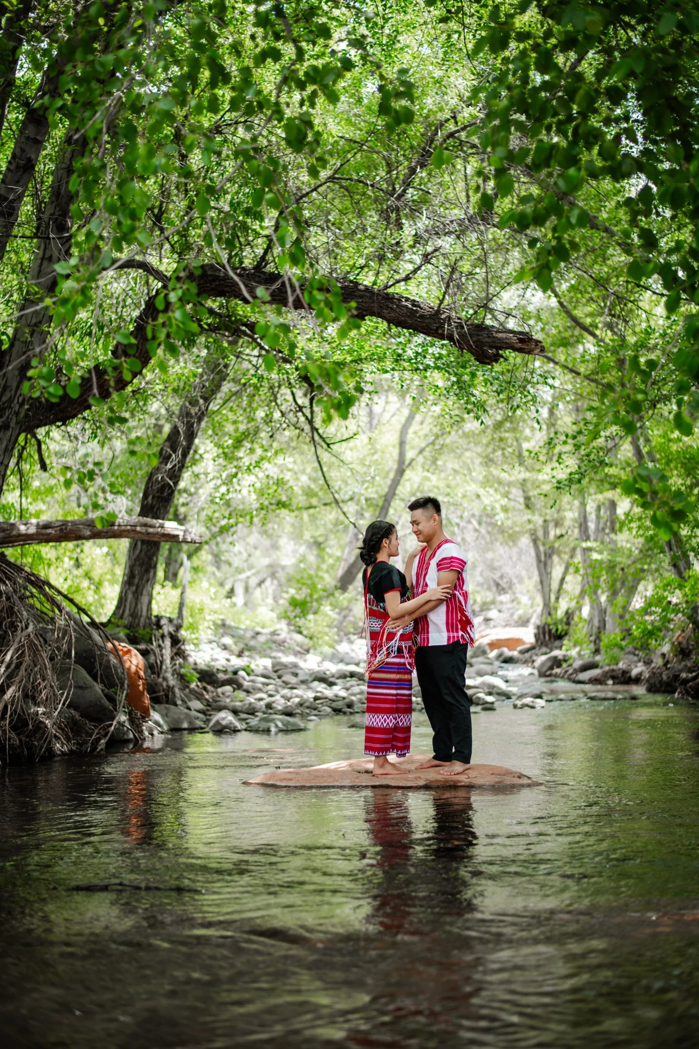 A couple standing on a large rock in the middle of a shallow stream, surrounded by lush green trees, under a canopy of green leaves, with soft sunlight filtering through.