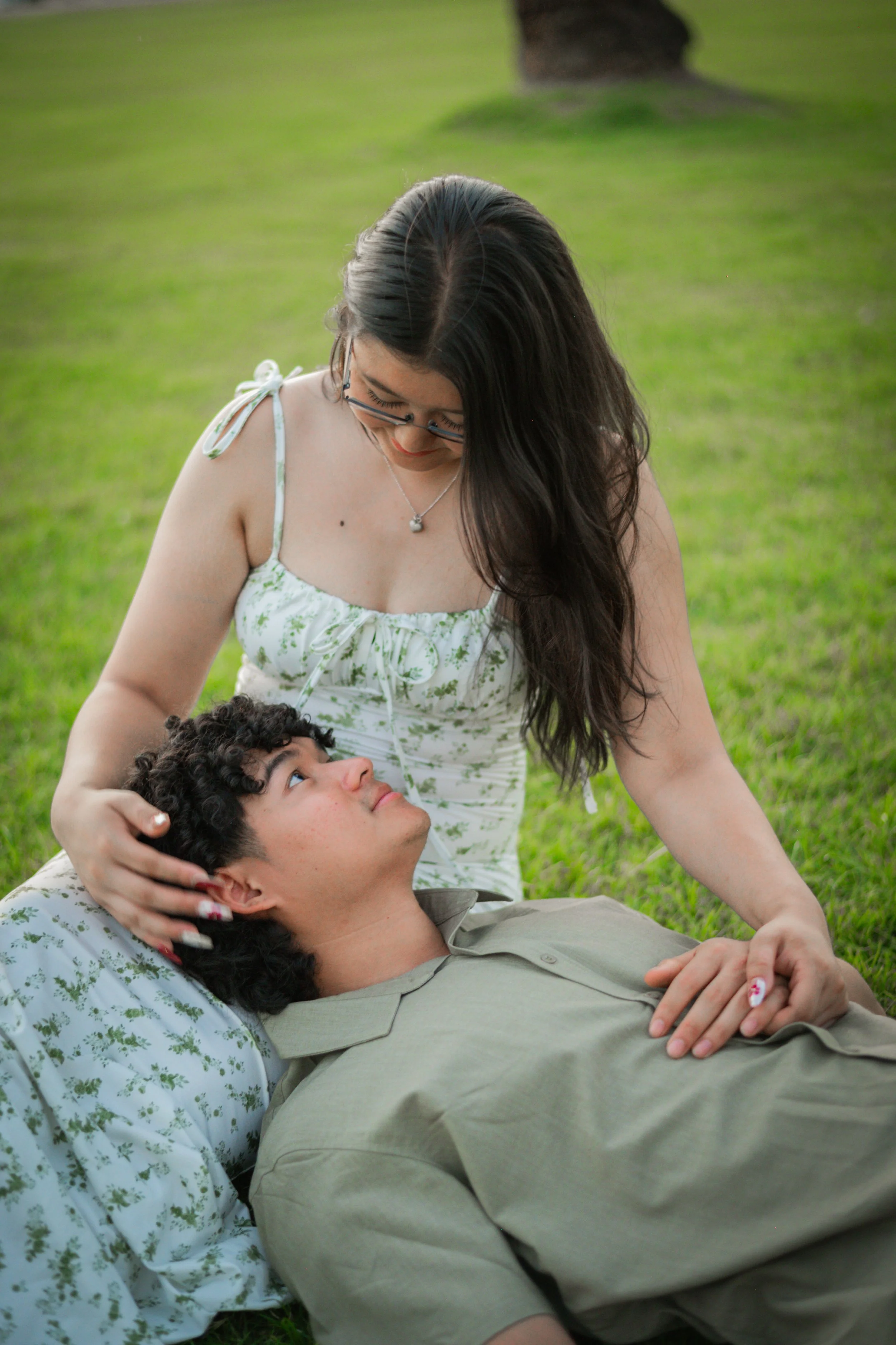 A young woman with long dark hair, glasses, and a white floral dress looks down affectionately at a young man with curly hair, lying on the grass. The woman has her hand on the man's head, and they are outdoors on a lush green lawn.