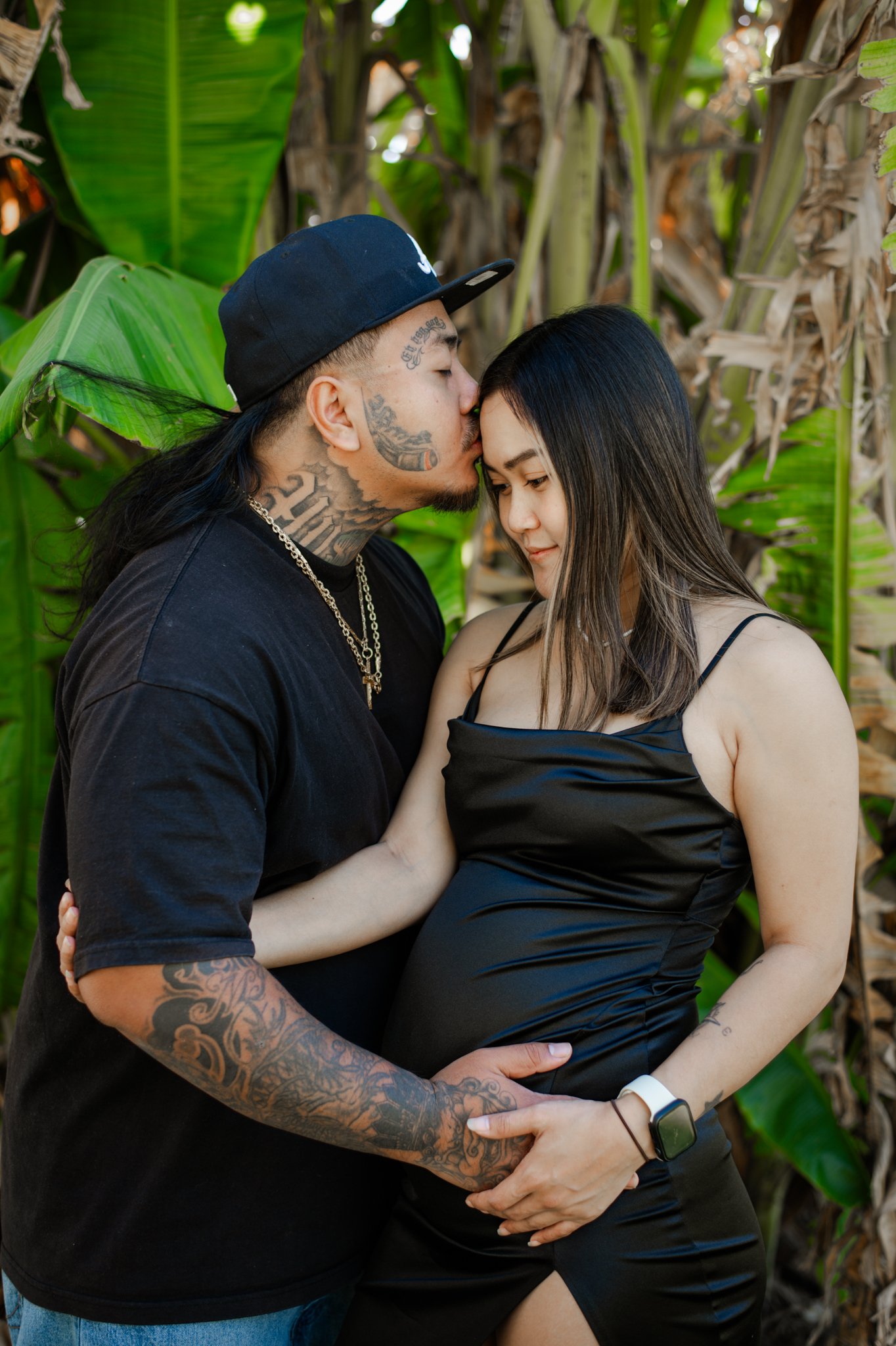 A couple embraces outdoors, with the man kissing the woman's forehead and the woman smiling softly, surrounded by large green tropical leaves.