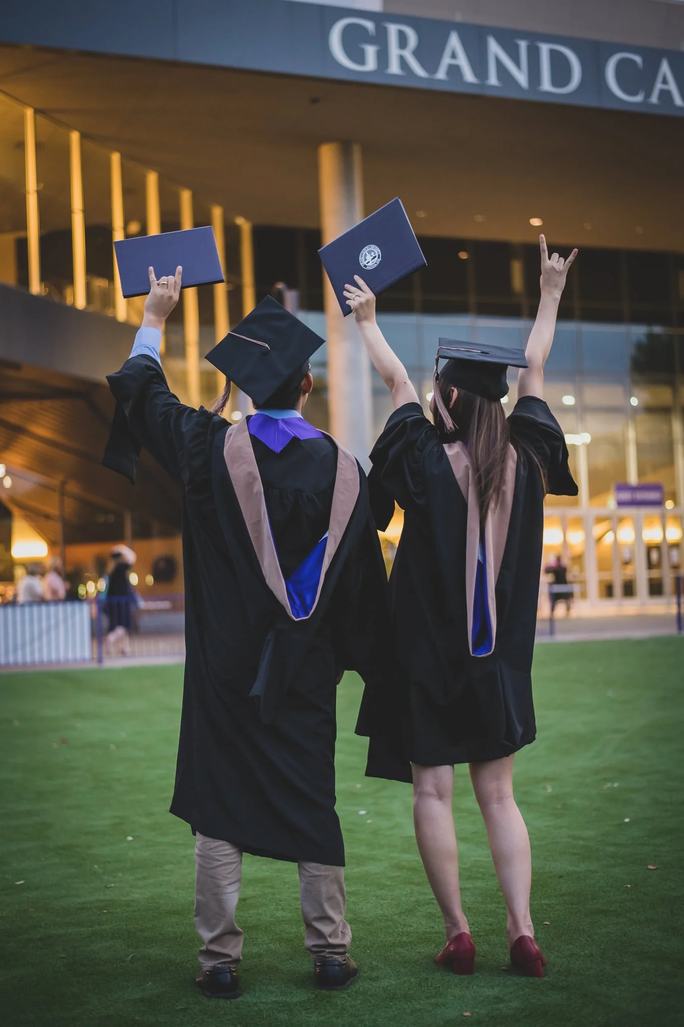 A group of graduates in caps and gowns celebrating at a graduation ceremony outside a modern building, with some holding diplomas in the air and one making a rock and roll sign with her hand.