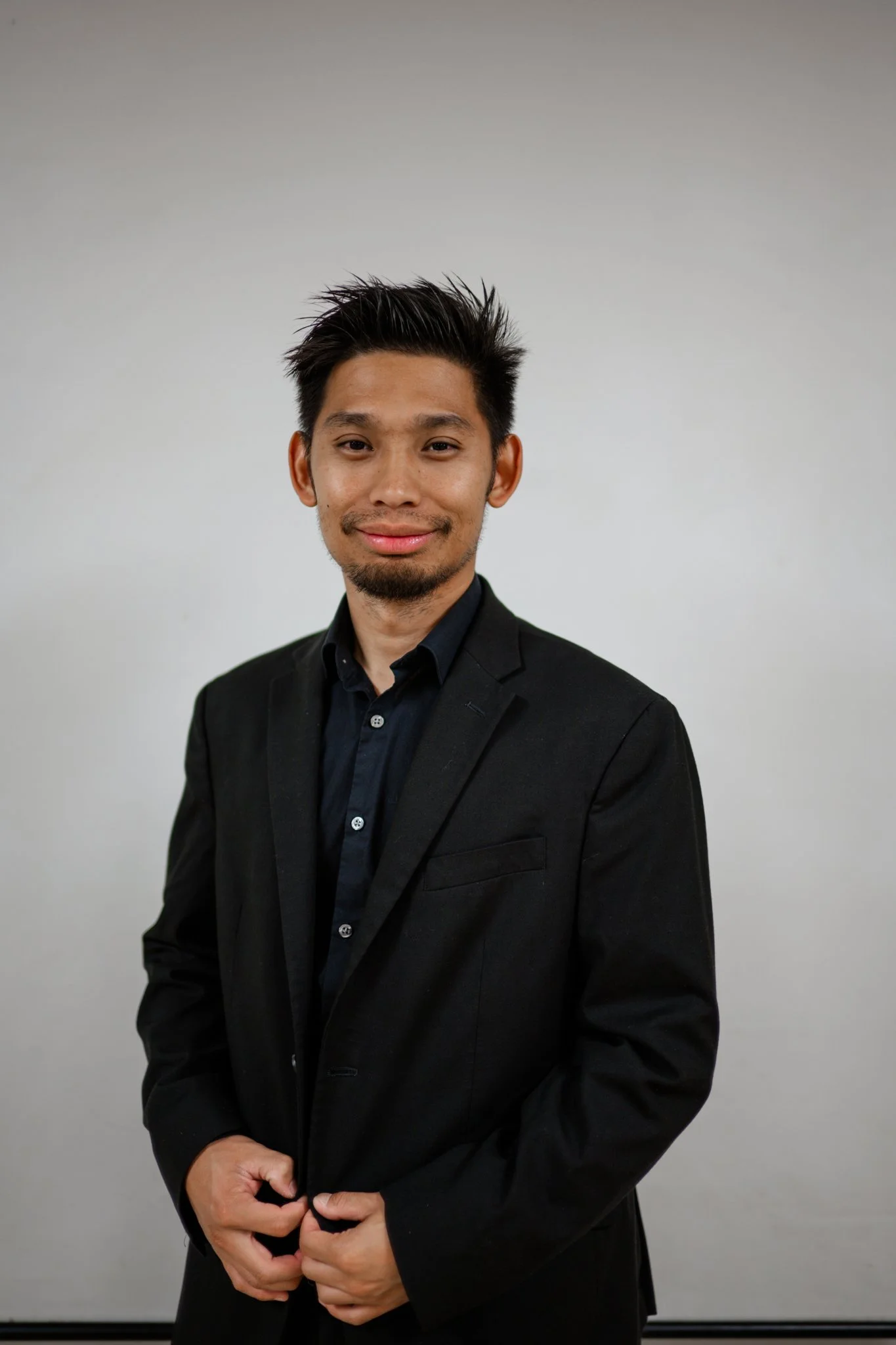 A young man with dark hair, wearing a black suit and black shirt, standing in front of a plain gray wall, smiling slightly.