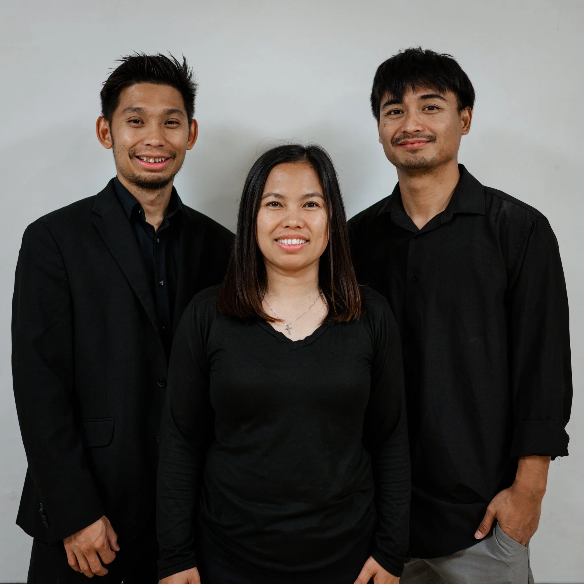 Three people standing together in front of a plain white wall, all dressed in black shirts, smiling at the camera.