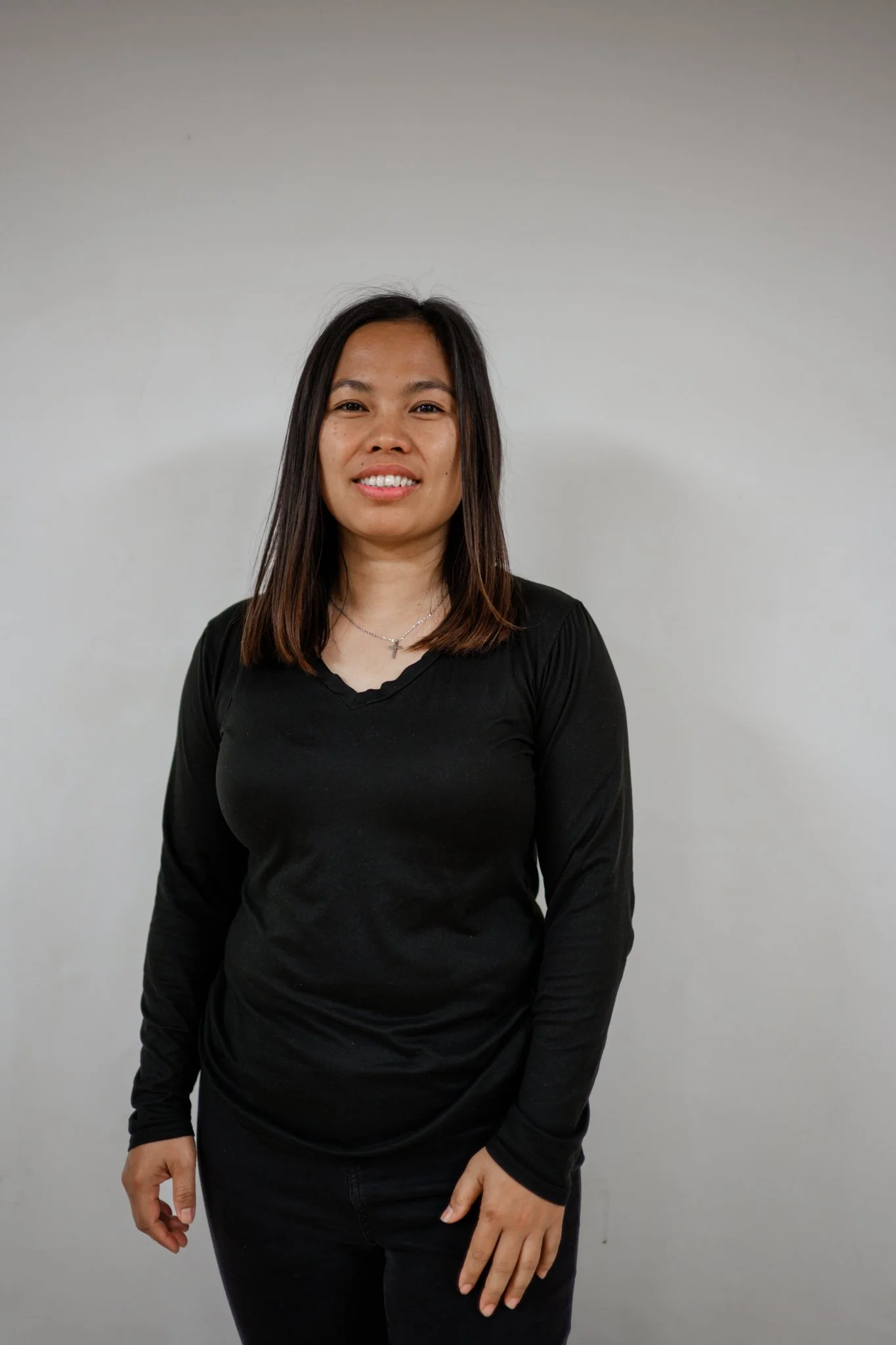 A woman with shoulder-length dark hair standing against a plain light-colored wall, wearing a black long-sleeve shirt and black pants, smiling gently.