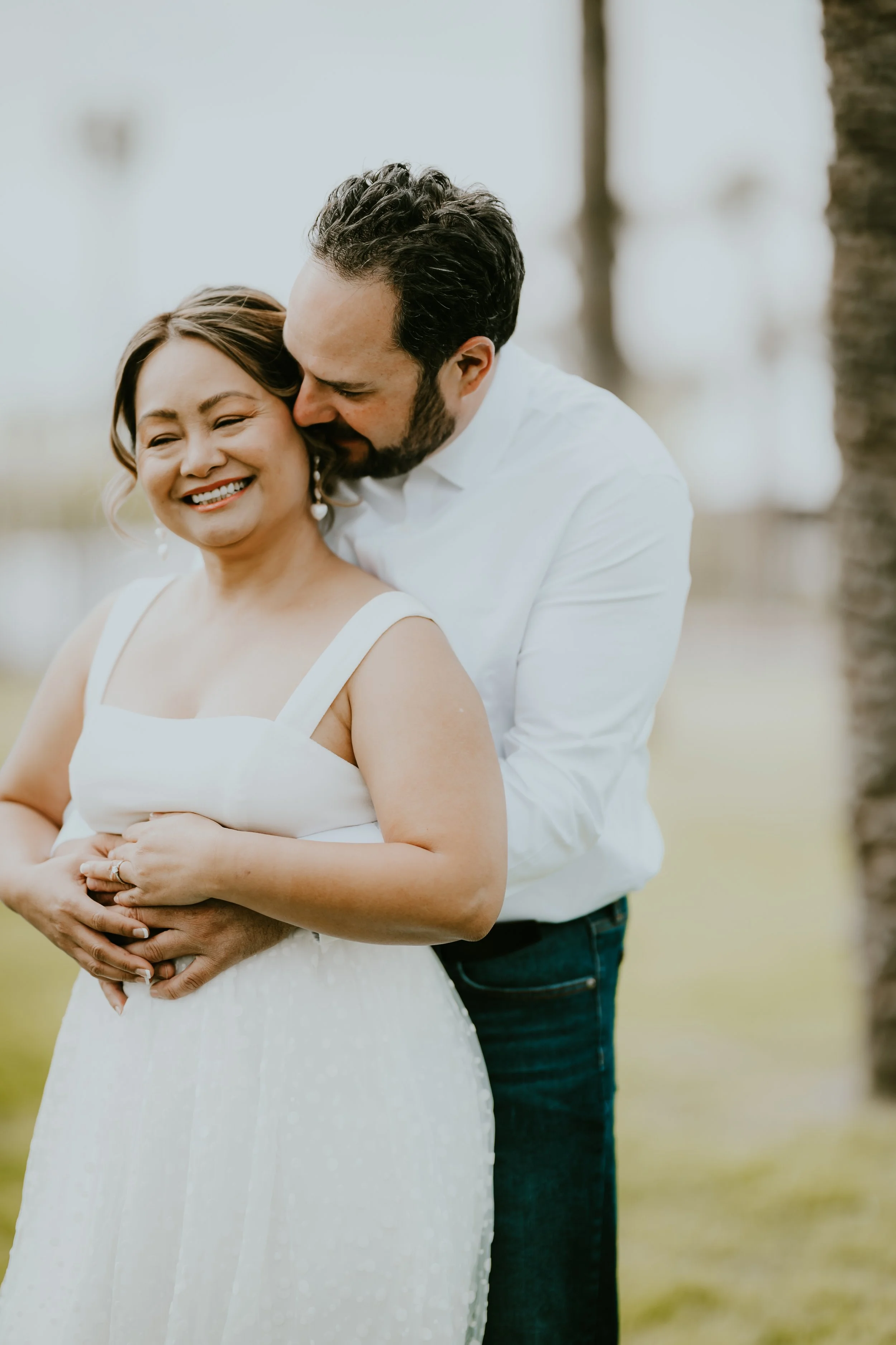 A couple embracing outdoors, with the man kissing the woman on her cheek. She is smiling, wearing a white dress and earrings, while he is dressed in a white shirt and blue jeans. The background is blurred with trees and a stone wall.