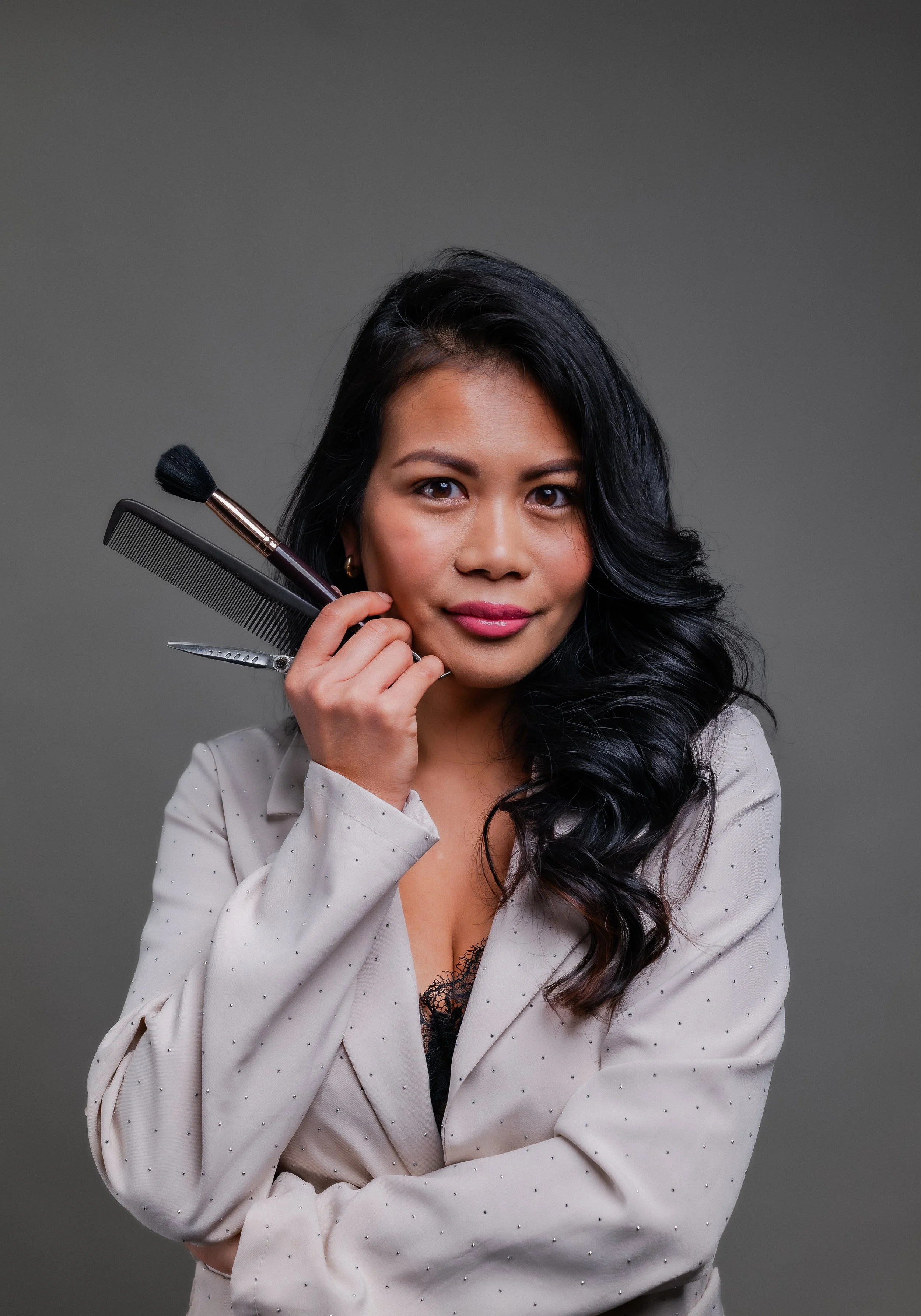 Eine Frau mit dunklen, gewellten Haaren, die ein Kamm, eine Nagelschere und einen Make-up-Pinsel hält, vor grauem Hintergrund, trägt eine weiße Jacke mit kleinen schwarzen Punkten.