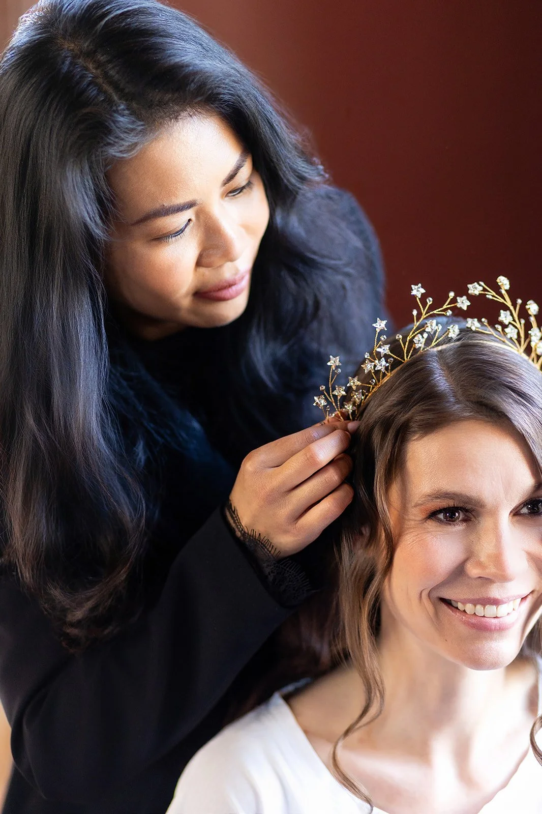 Eine Braut mit braunen, lockigen Haaren trägt eine goldene Krone mit kleinen Blumen, während Eve Chaimongkol mit schwarzen Haaren und einem schwarzen Oberteil sie sanft an der Krone berührt, beide lächeln.