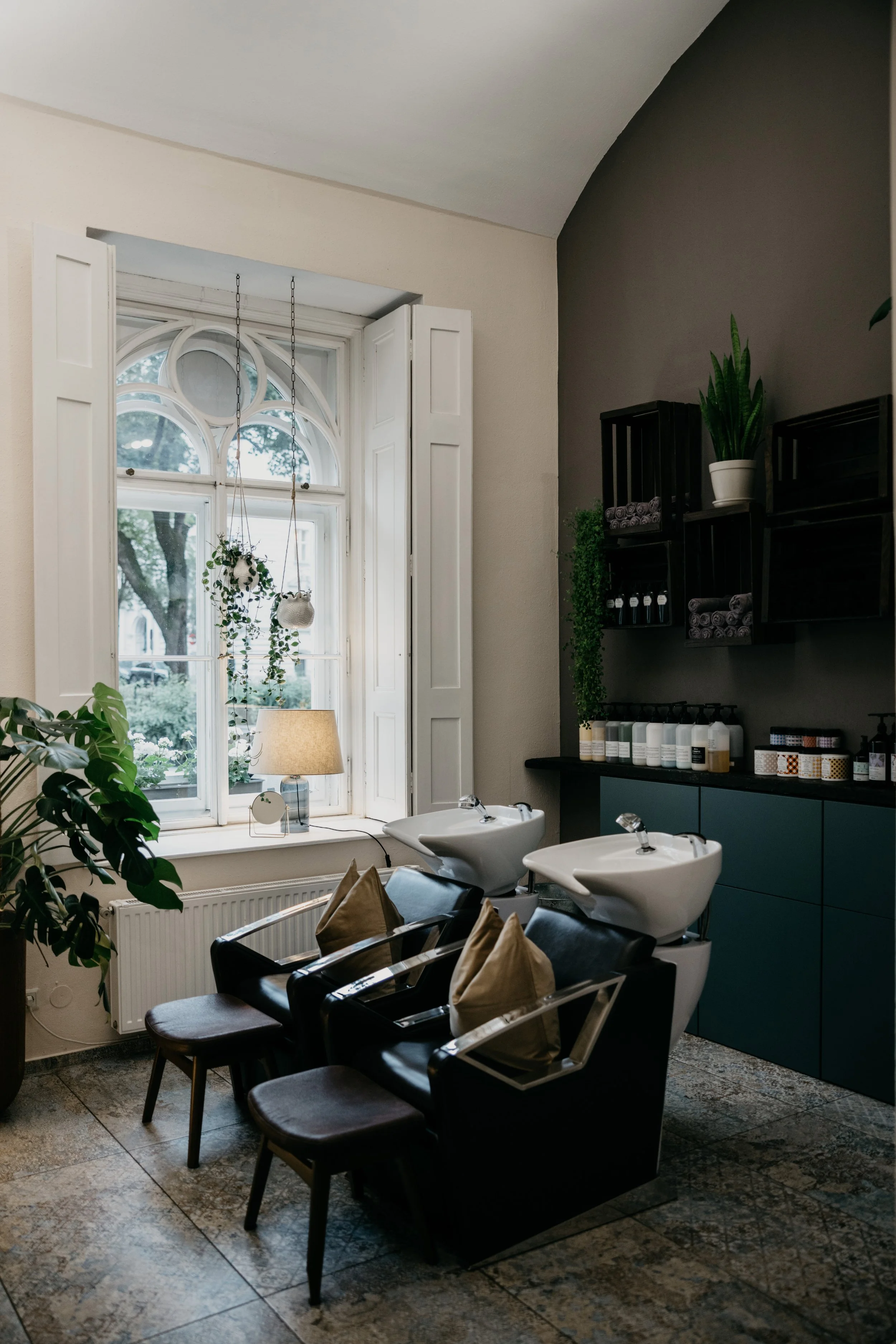 Innenraum eines Friseursalons mit Waschsitzen, schwarzem Stühlen, Pflanzen, großen Fenstern und einem Regal mit Pflegeprodukten.