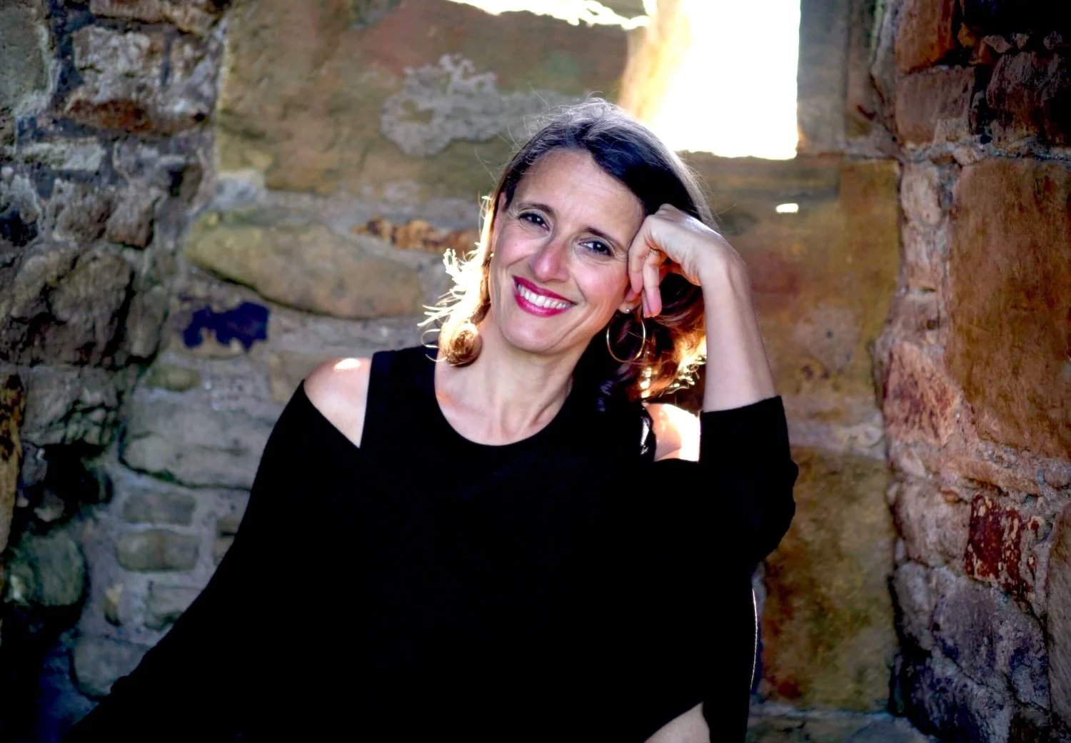 A woman with shoulder-length brown hair and hoop earrings, smiling and resting her head on her hand, sitting in a stone room with sunlight shining through a small window behind her.