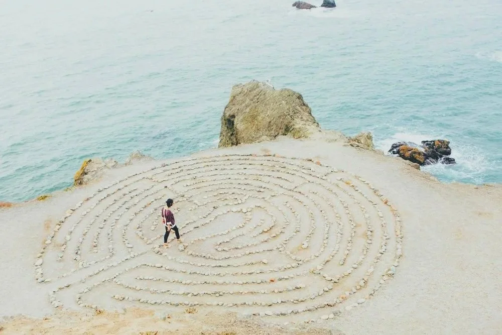A person walking through a spiral stone labyrinth on a coastal cliff with the ocean in the background.