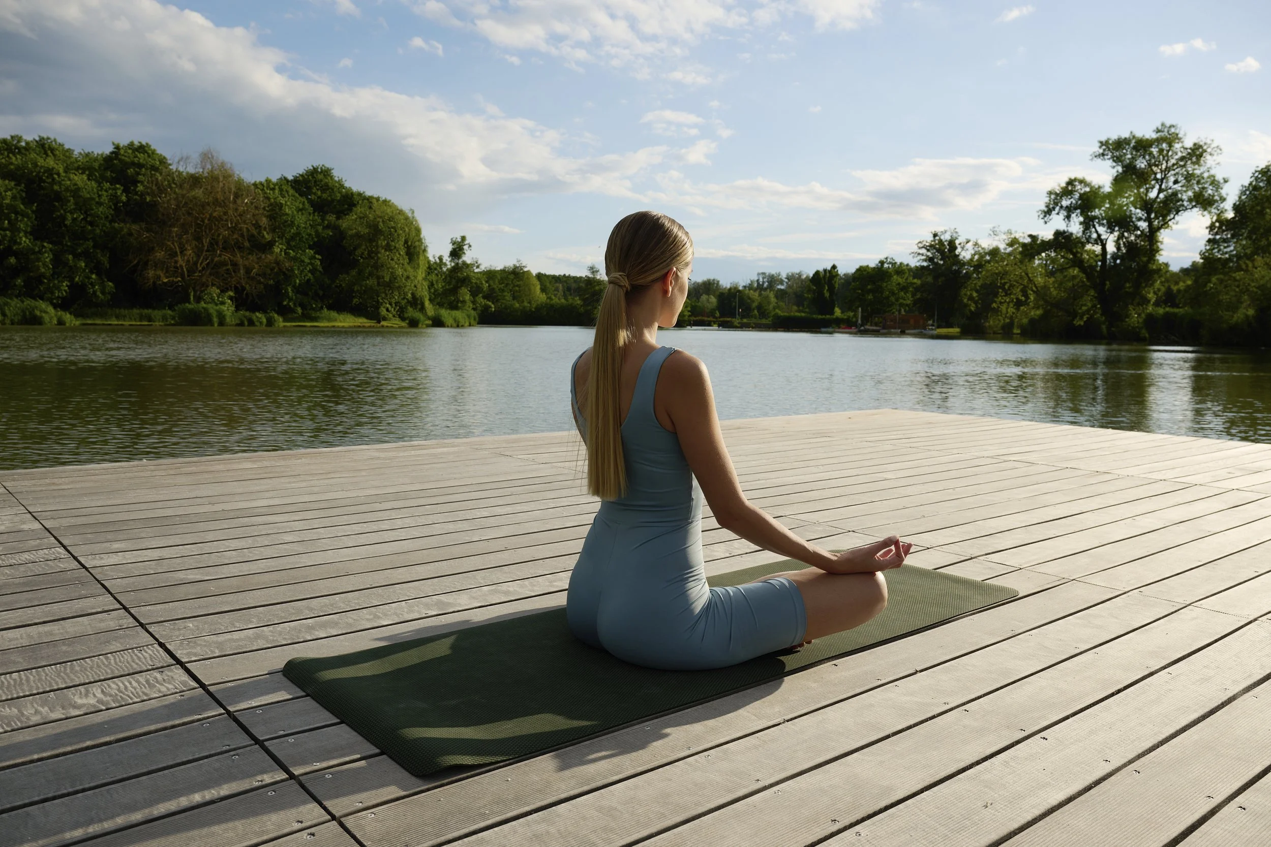 A woman practicing yoga on a mat by a lake, sitting in a lotus pose with her hands in a mudra, surrounded by trees and a partly cloudy sky.