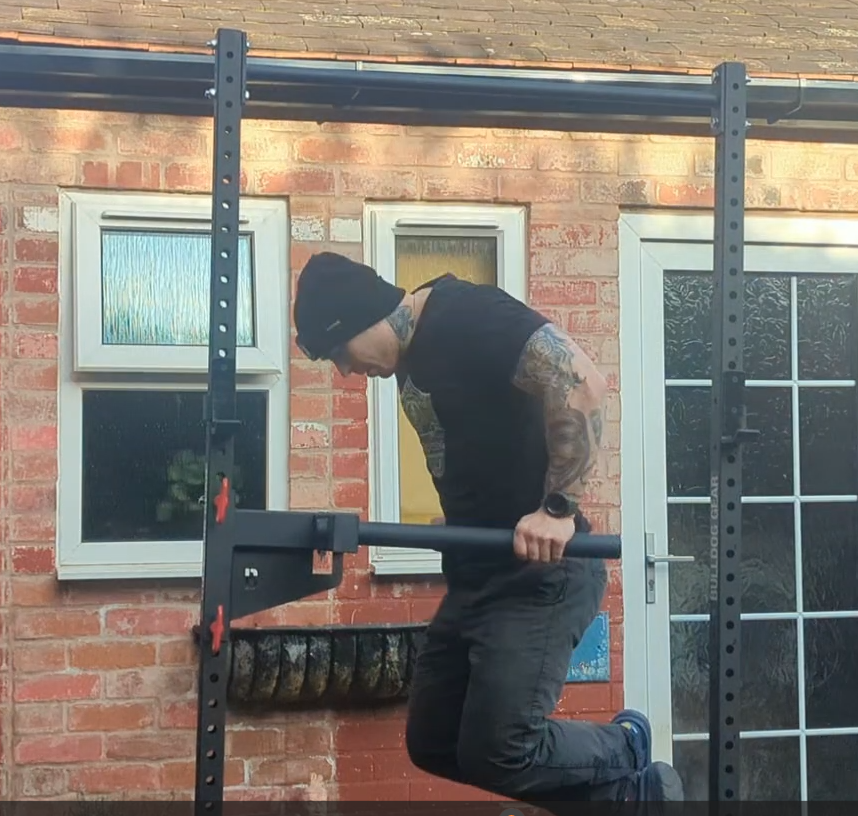 A man with tattoos, wearing a black beanie and black clothes, is hanging onto a pull-up bar outside a brick house.