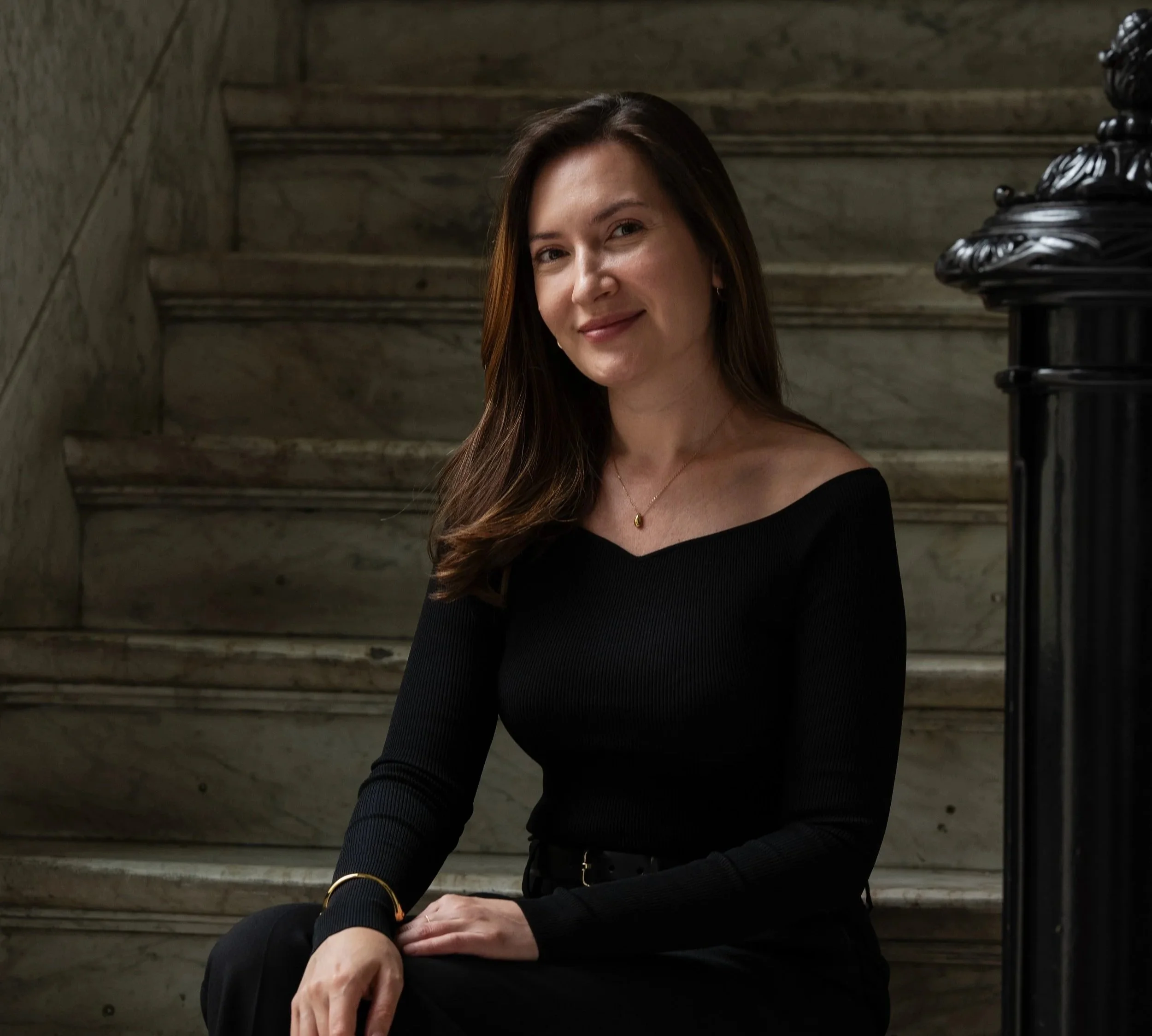 A woman with long brown hair wearing a black off-shoulder top and black pants, sitting on marble stairs indoors.