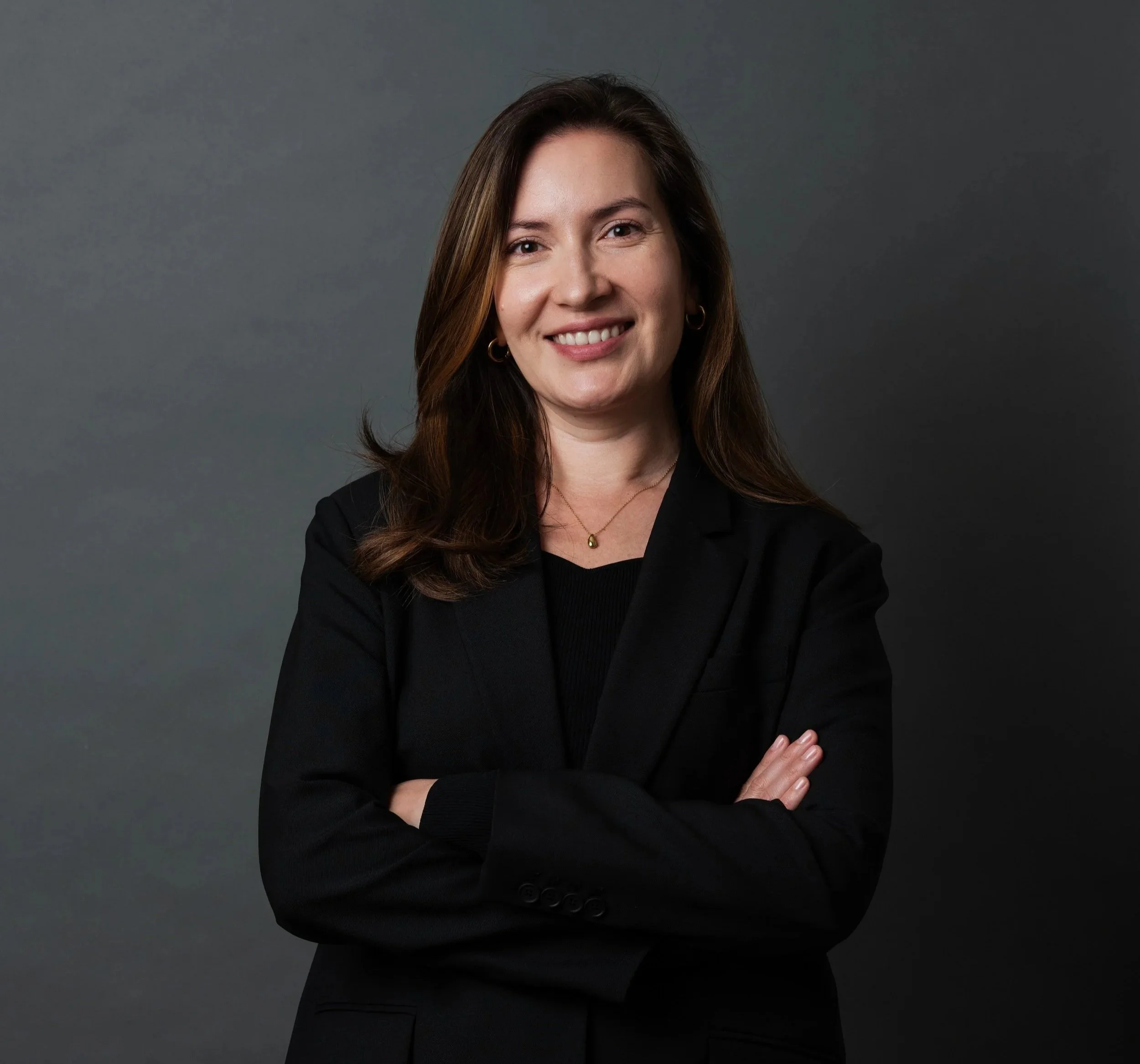 A professional woman with long brown hair wearing a black blazer and top, smiling with arms crossed, standing against a dark gray background.