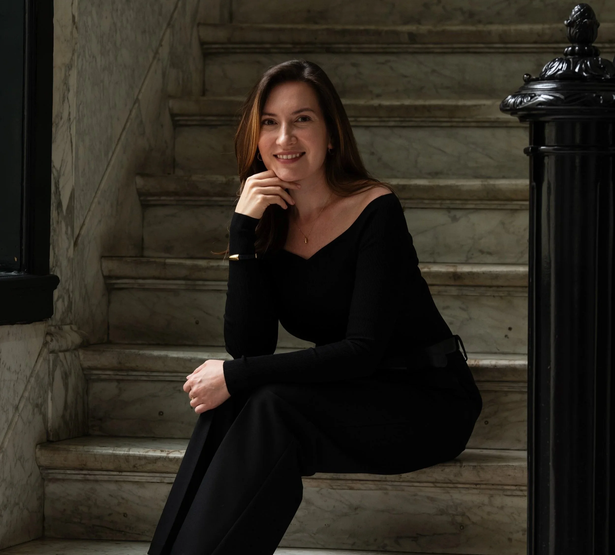 A woman with long brown hair sitting on marble stairs, smiling at the camera, wearing a black long-sleeve top and black pants, with her left hand resting on her knee and her right hand touching her chin.