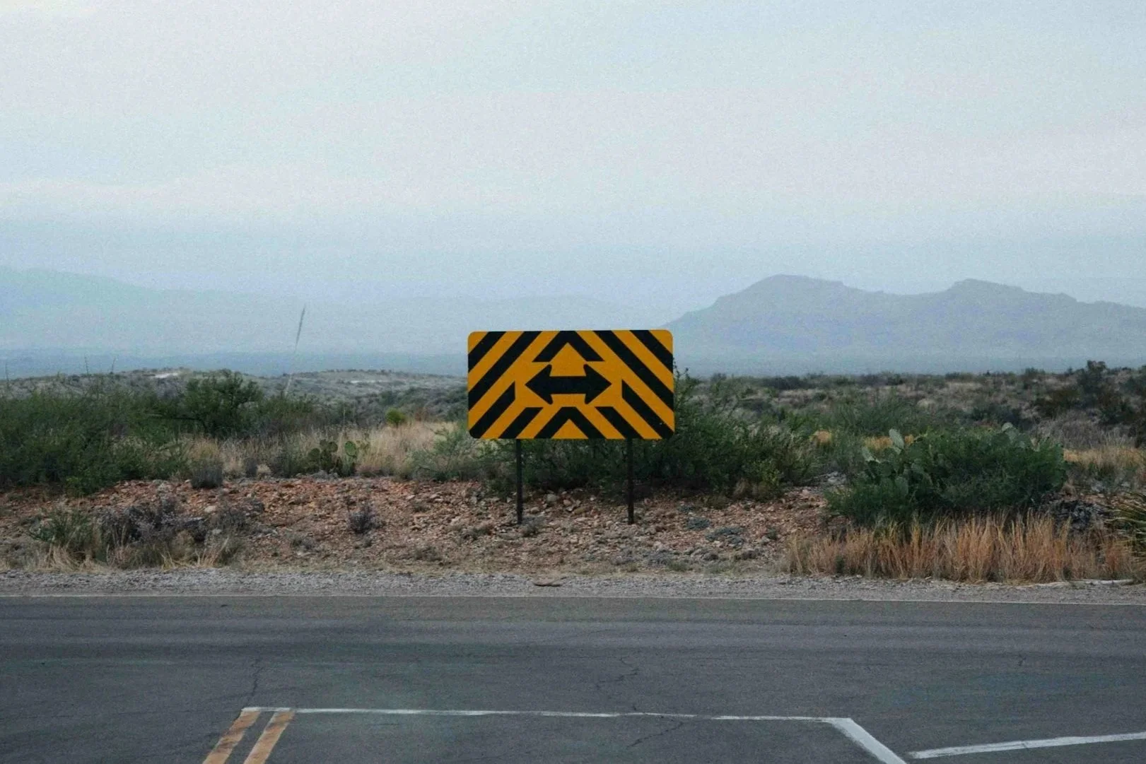 A roadside warning sign with black arrows pointing left and right on a yellow and black striped background, in a desert landscape with sparse vegetation and mountains in the distance.