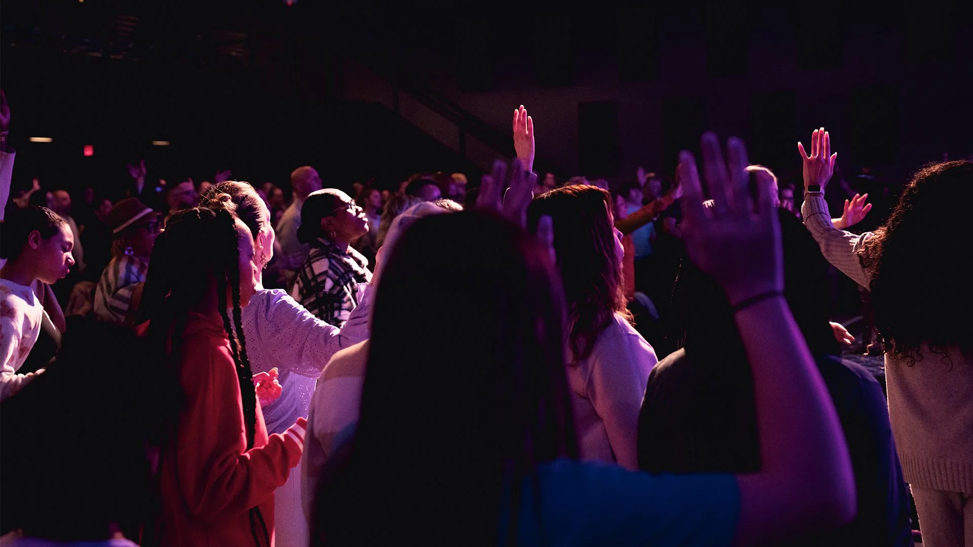 A crowd of people in a dark auditorium with purple lighting, with some raising their hands.