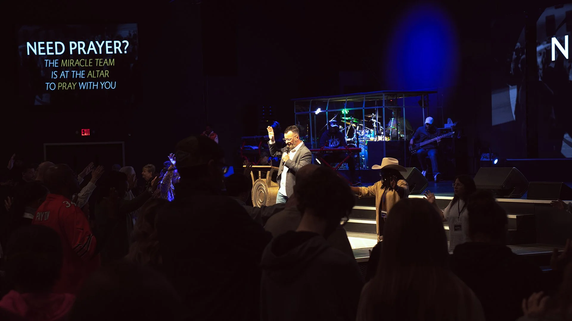 A man speaking into a microphone on stage while congregation members raise their hands during a church service or prayer event. The stage has musical instruments, and a large screen displays text about prayer and the miracle team.