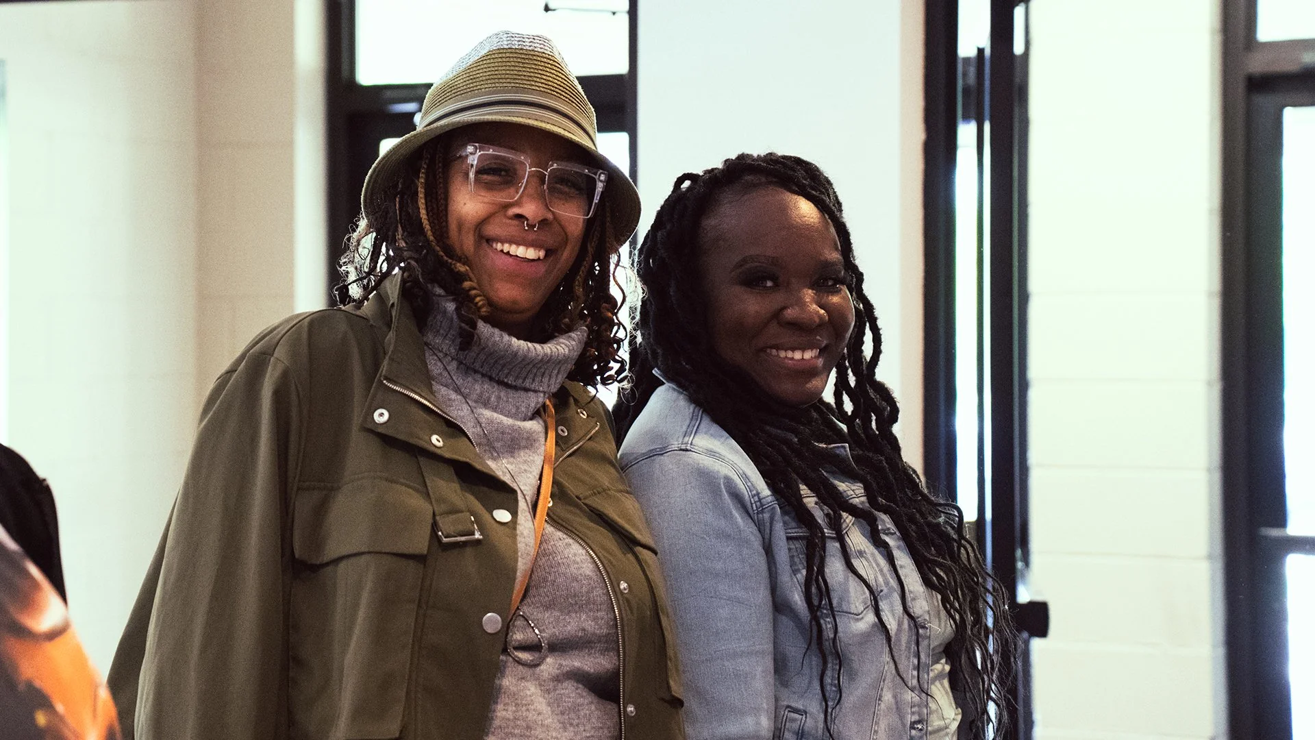 Two smiling women standing indoors near a window, one wearing a hat and glasses, the other with long twists in her hair.
