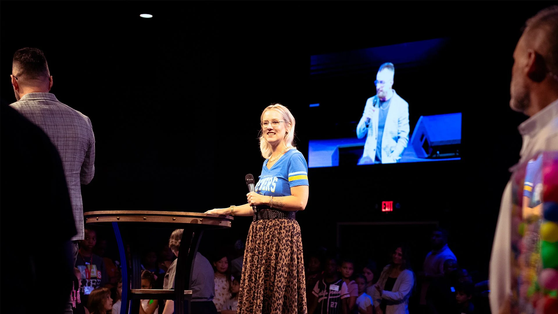 A woman standing at a high table and speaking into a microphone in a dark room, with a large screen behind her displaying her image, while several people watch in the background.