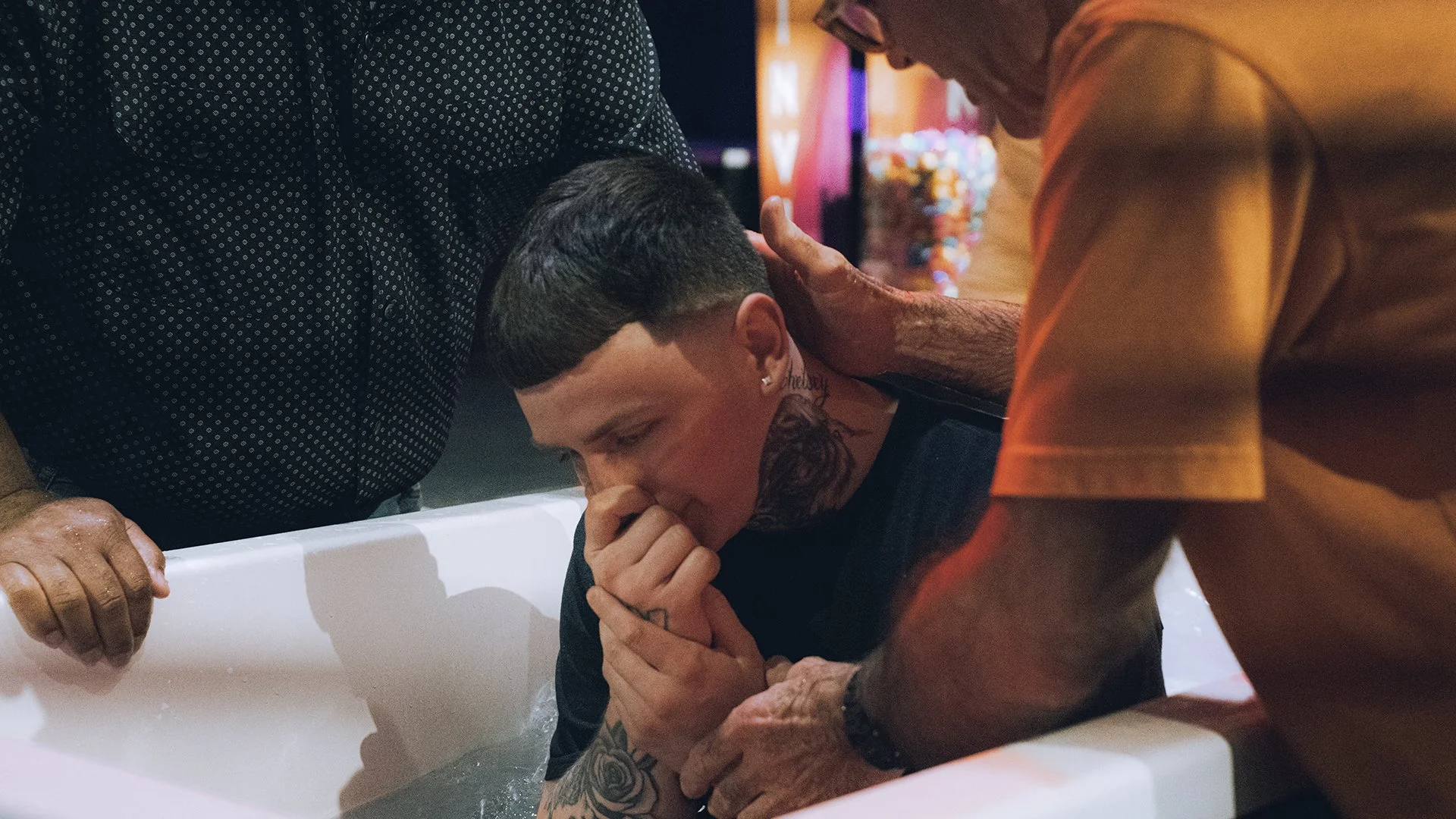 A young man with tattoos on his neck and hands is praying with his eyes closed, while an older man in an orange shirt is placing a hand on his head in a baptism ceremony in a church or similar setting.