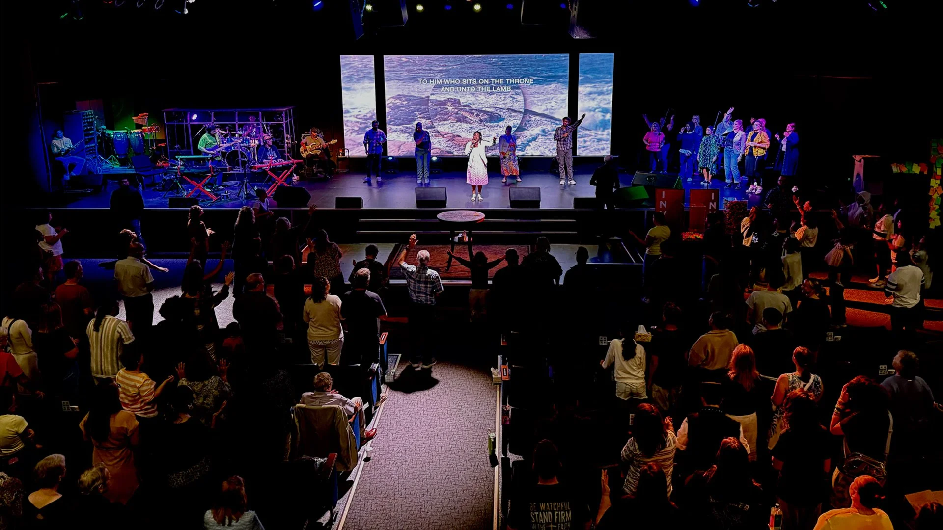 A large group of people gathered inside a church with a stage at the front where musicians and singers are performing. The audience is standing with some raising their hands. On the stage, there are musicians playing instruments and a screen behind them displaying lyrics.