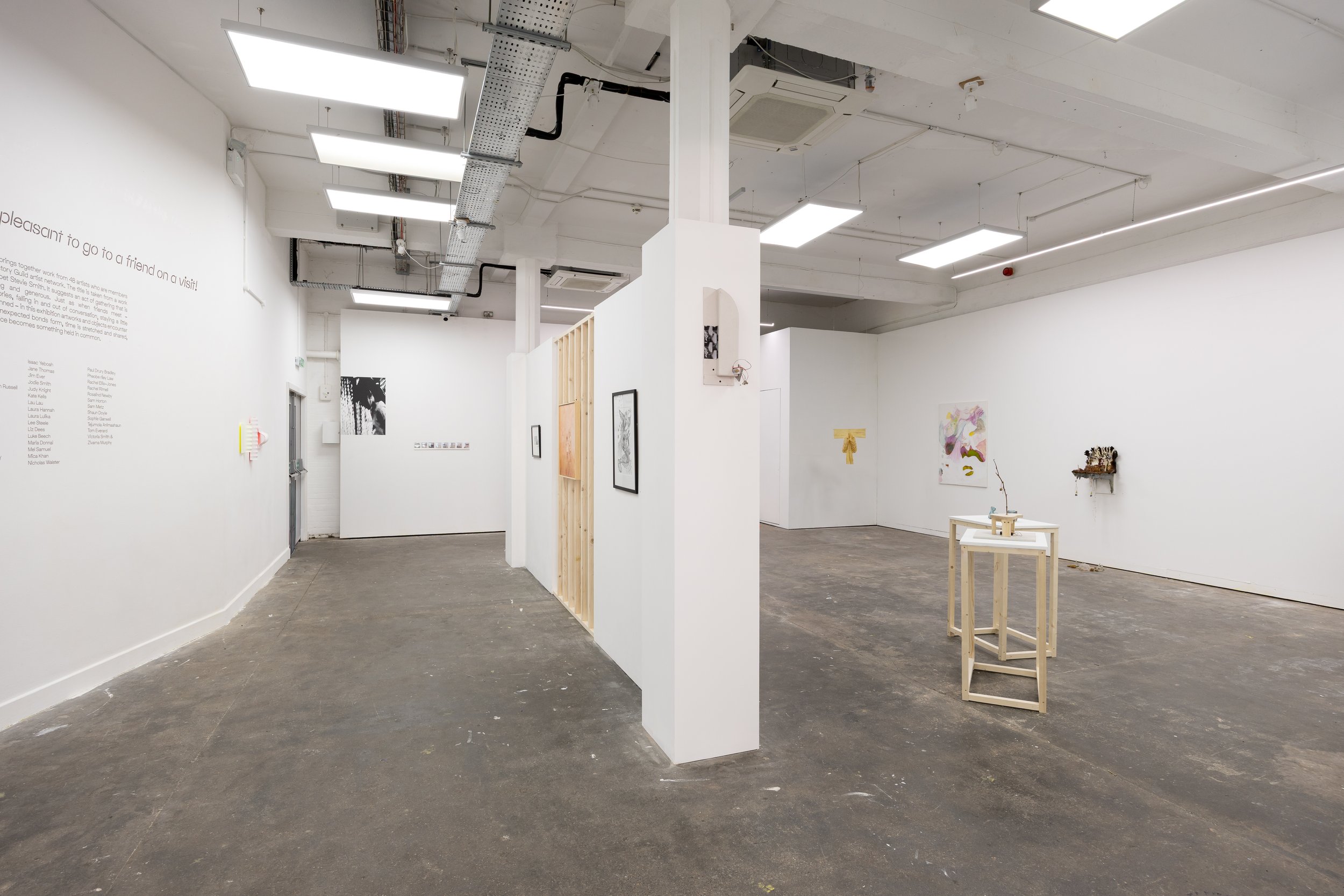 An art gallery with white walls, featuring framed artwork and wall text, with a concrete floor and a partial view of a wooden display stand.