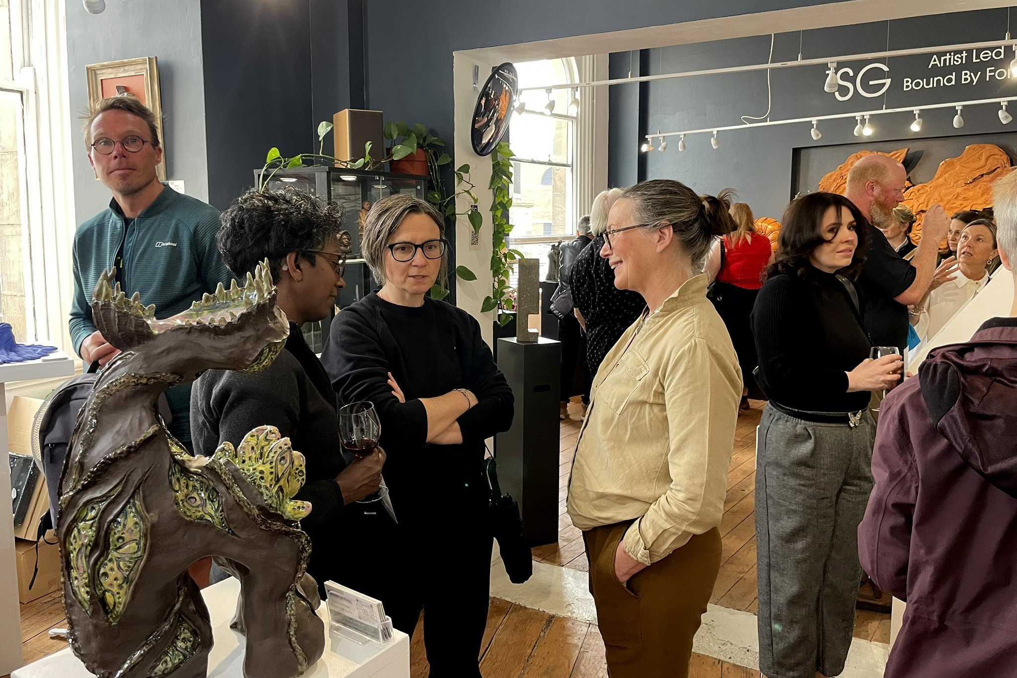 Gallery event with people engaging in conversation. In the foreground, a woman holds a glass of red wine, and a man with glasses and gray hair is talking to her. There are multiple people in the background, some near artworks and displays, inside a gallery space with dark-colored walls, large windows, and track lighting.