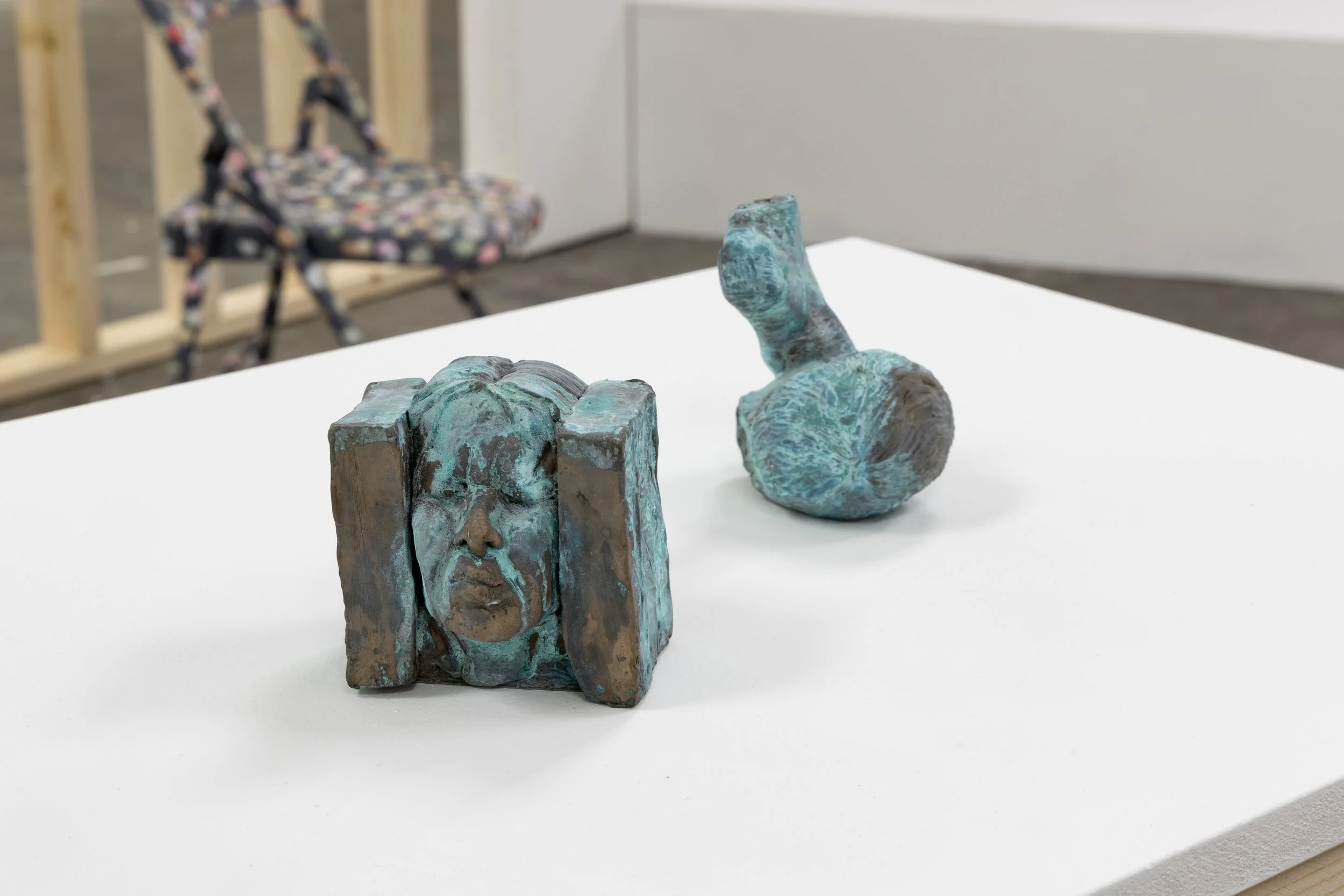 Two abstract sculptures, one resembling a face and the other a figure, are placed on a white table in an art gallery.
