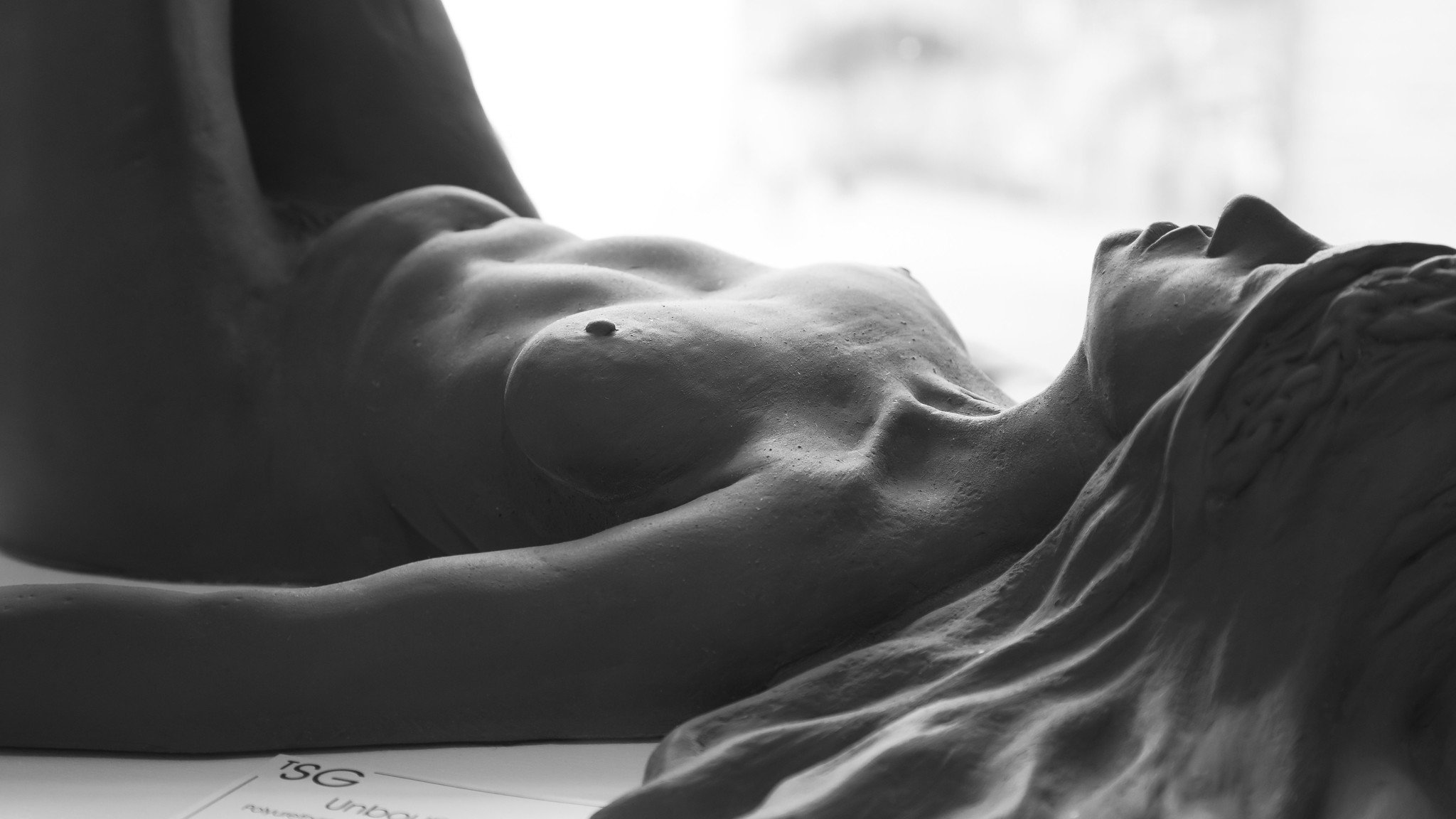 Close-up black and white photograph of reclining female nude sculpture with detailed facial features, lying on her back with her eyes closed.