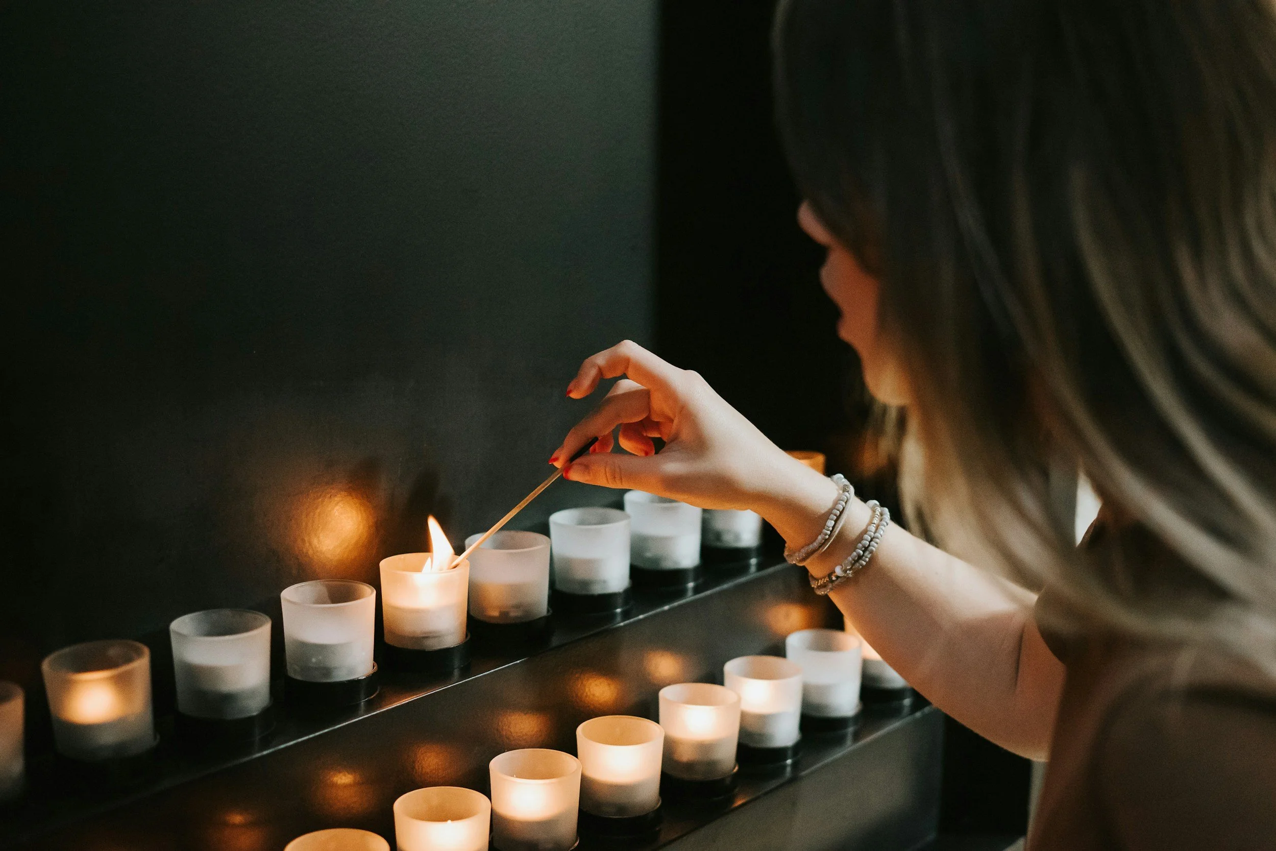 Woman lighting a candle with a matchstick in a dark room, surrounded by row of lit candles.