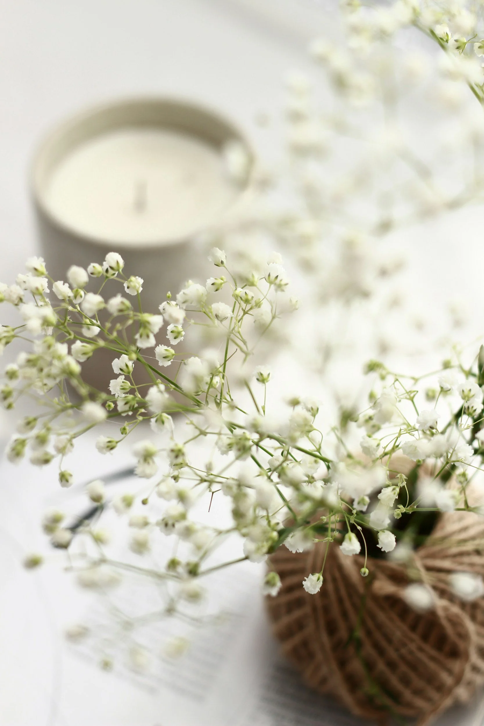 A bouquet of white baby's breath flowers wrapped with a ribbon, with a lit white candle in the background on a white surface.