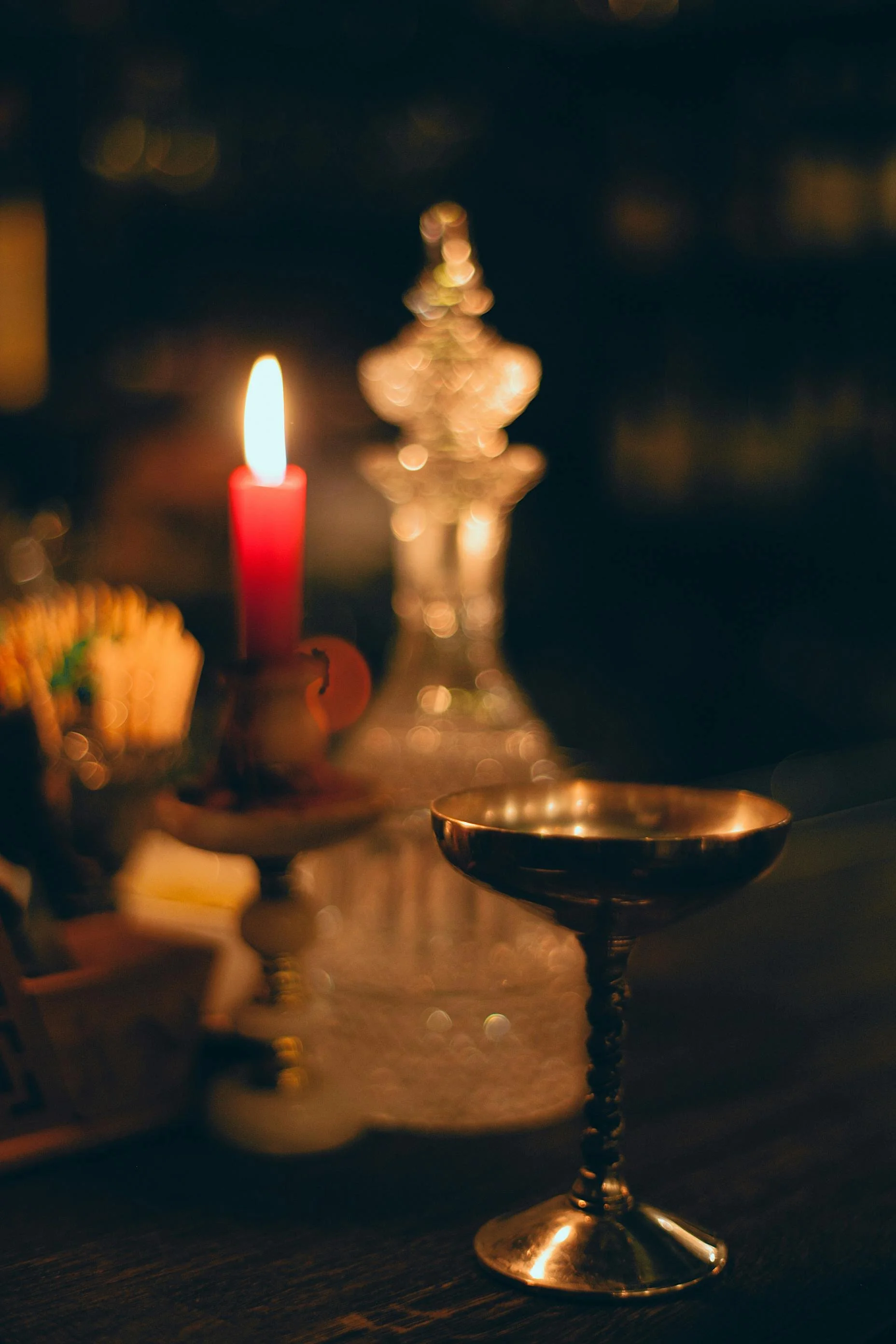 A dimly lit table featuring a red candle in a candle holder, a decorative glass object, and a metallic goblet, creating a warm, festive atmosphere.