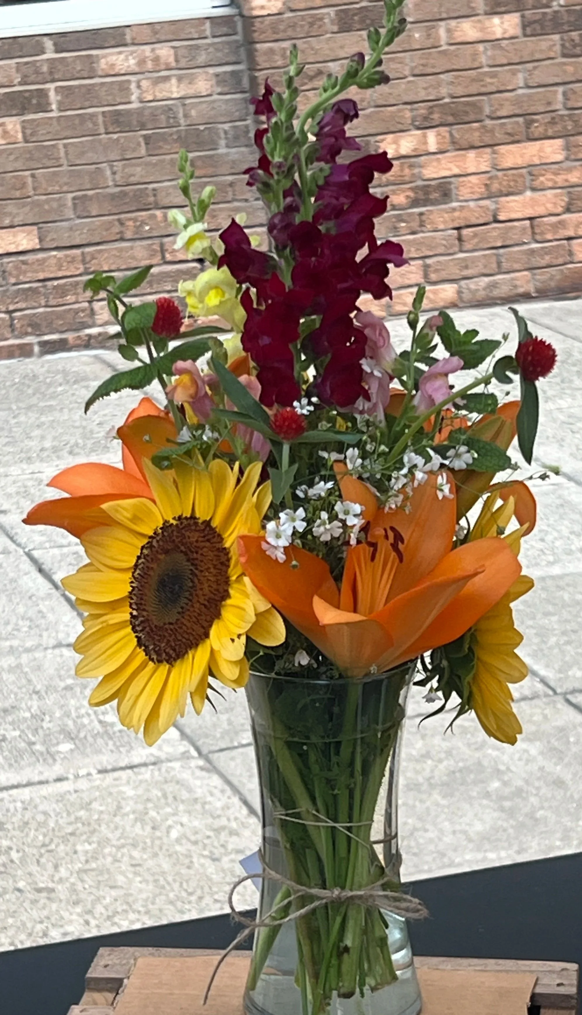 Colorful flower arrangement in a clear glass vase including sunflowers, lilies, snapdragons, and other small flowers, with a brick wall and concrete surface background.