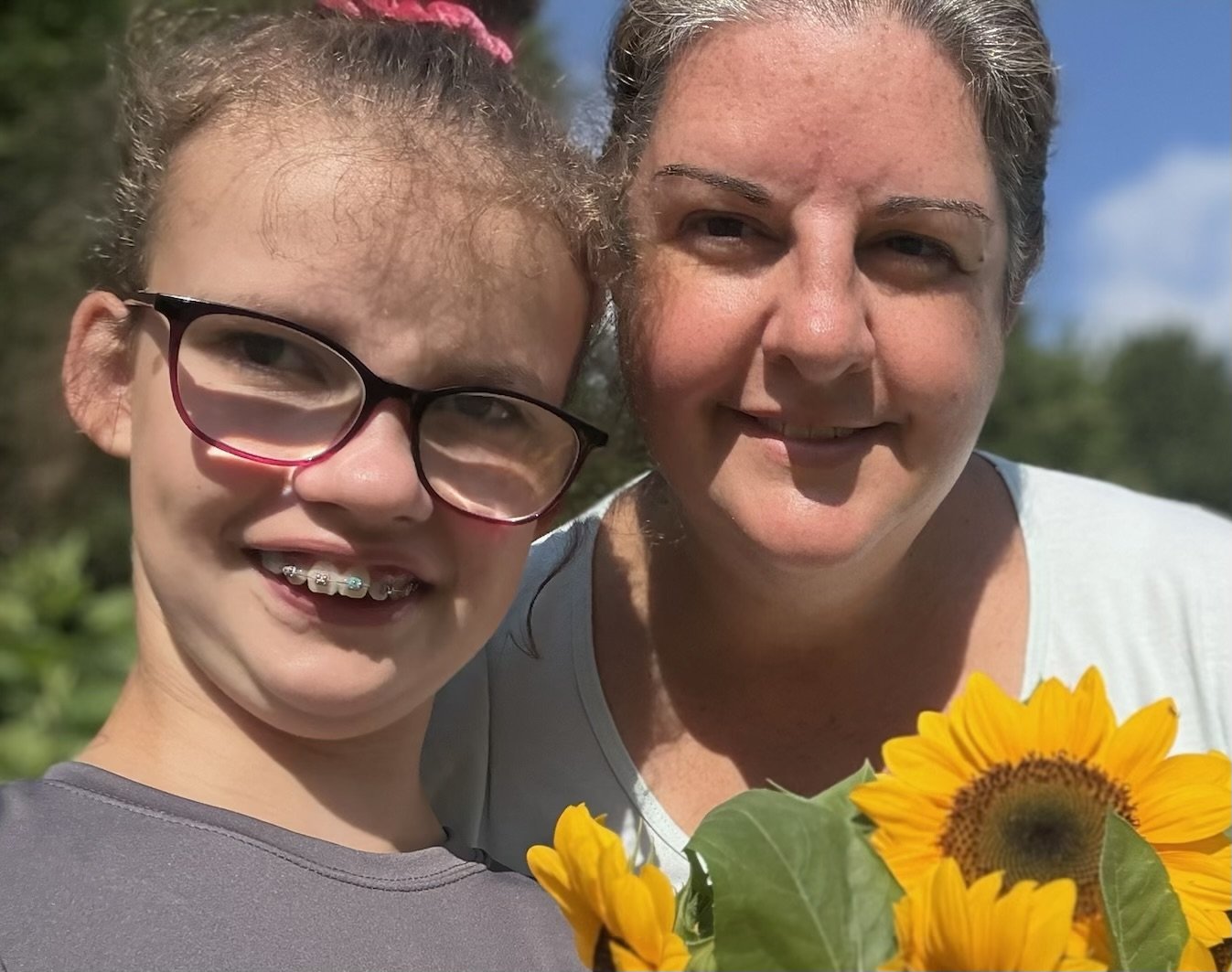 A young girl and an older woman standing outdoors, smiling for a selfie with bright sunflowers, the girl wearing glasses and braces, and the woman with short gray hair, under a clear blue sky.