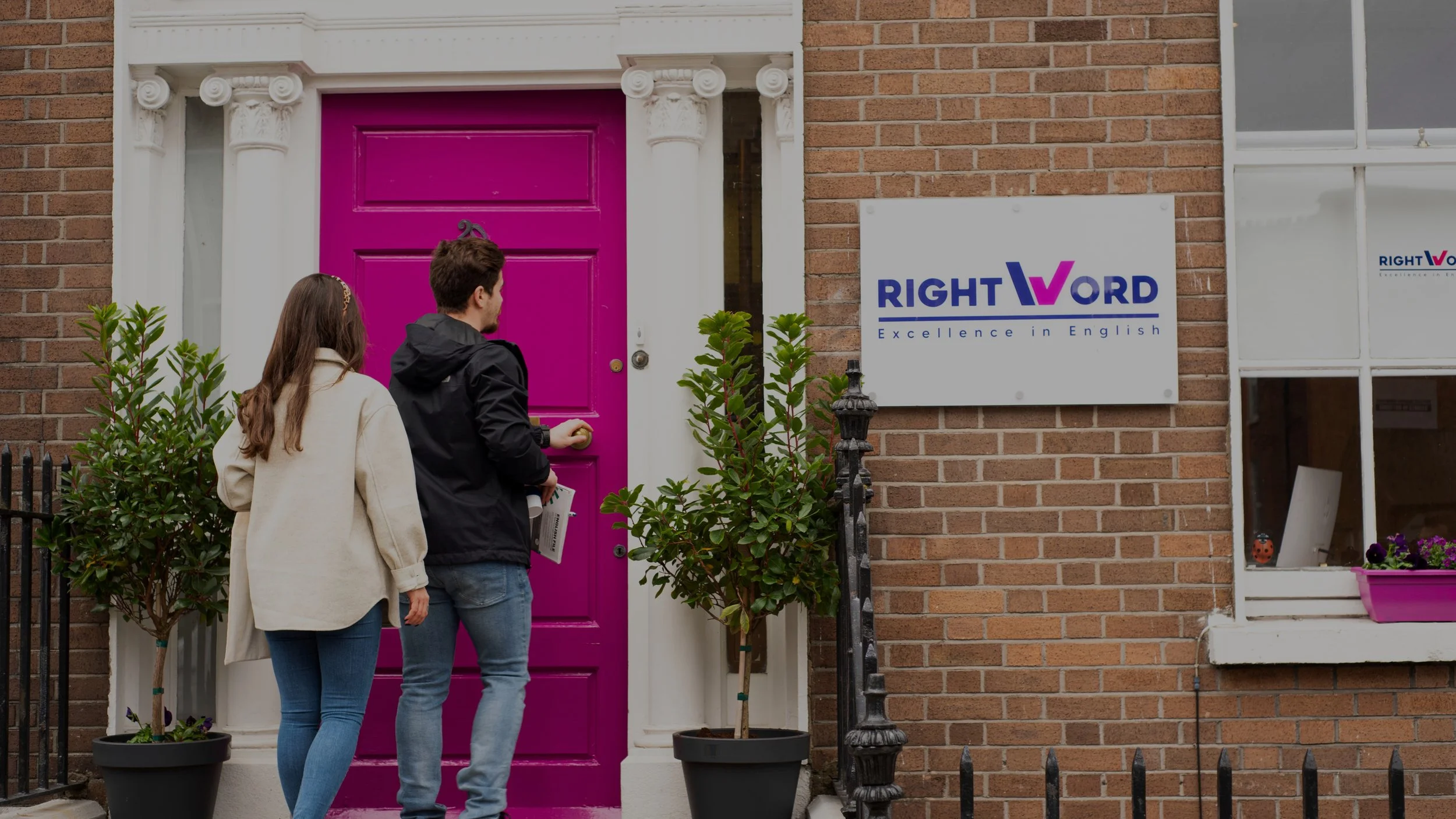 A man and a woman standing outside a brick building with a bright pink door. The man is reaching for the door handle, and the woman is holding papers. There are two potted plants on either side of the door, and a sign that reads 'RIGHT WORD Excellence in English' is on the wall.