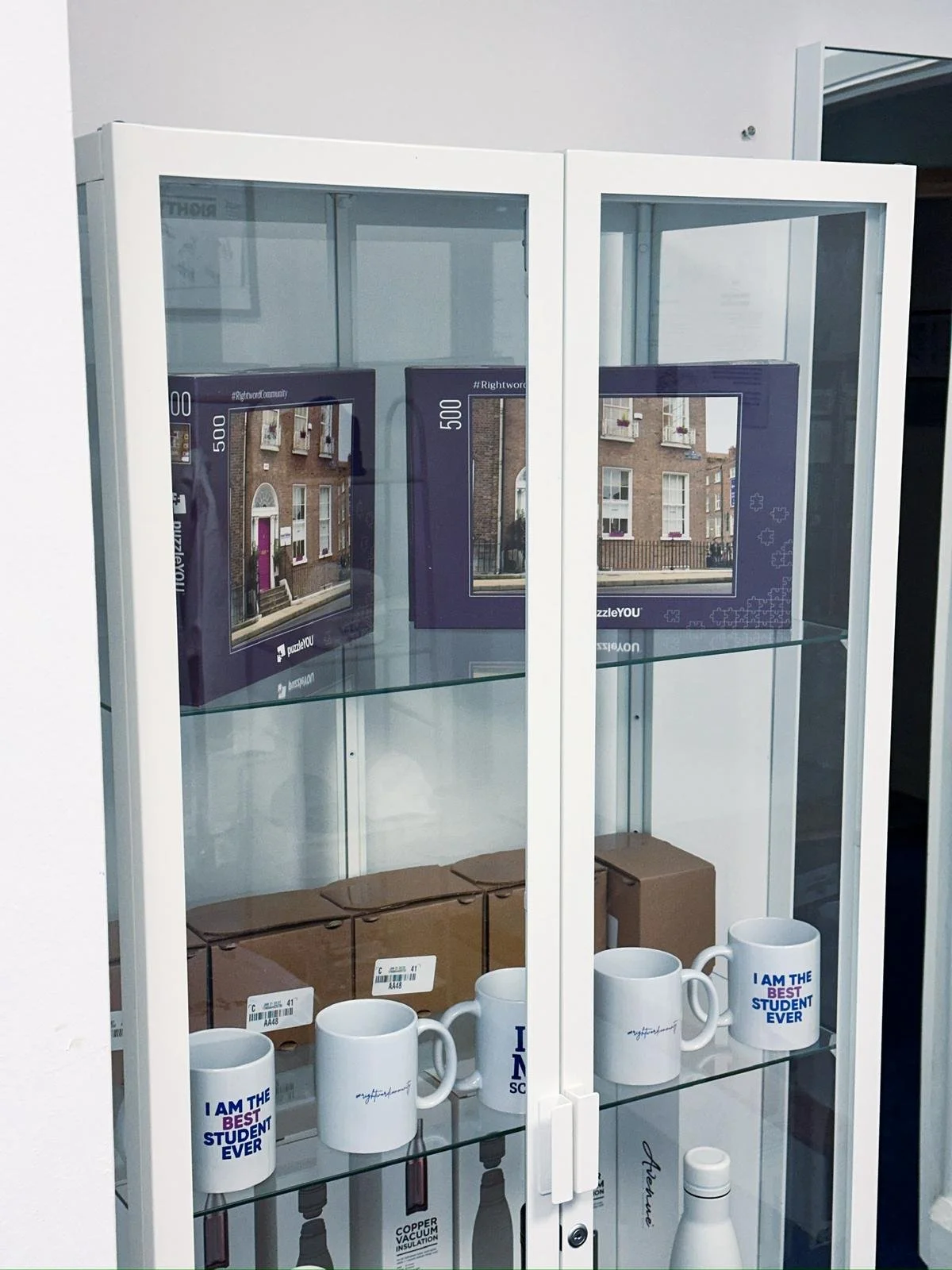 Display cabinet with ceramic mugs that say 'I am the best student ever,' and boxed puzzles with an image of a building, along with some brown boxes on the bottom shelf.