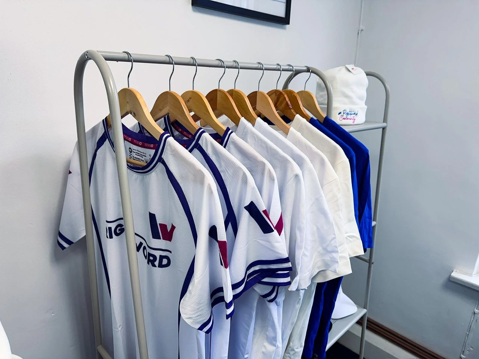 A clothing rack with white, blue, and white jerseys hanging on wooden hangers and a white towel with embroidery resting on the top shelf.