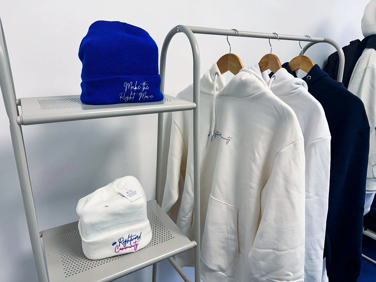 White and navy hoodies hanging on a rack, and two beanies on a metal shelf, one blue and one white, with embroidered text reading 'Make the Right Move' and 'Rightward Community' respectively.