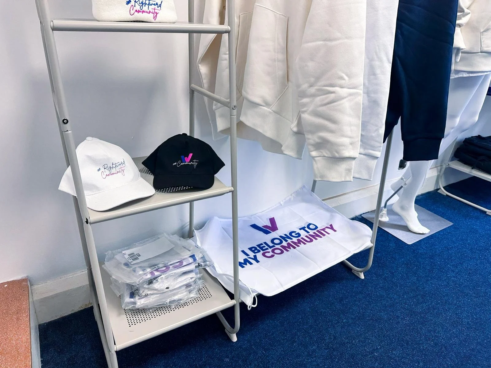 Display of clothing and accessories on a metal rack and shelf promoting community involvement; includes white and black caps, face masks, and a cloth with the phrase 'I BELONG TO MY COMMUNITY'.