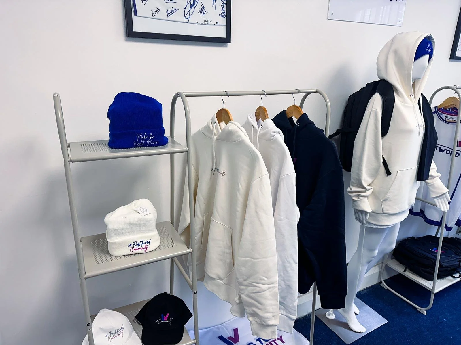 Display of clothing items including white, black, and navy hoodies on a rack, a mannequin wearing a white hoodie and black backpack, with hats and beanies on shelves, all branded with 'Rightward Community' logos.